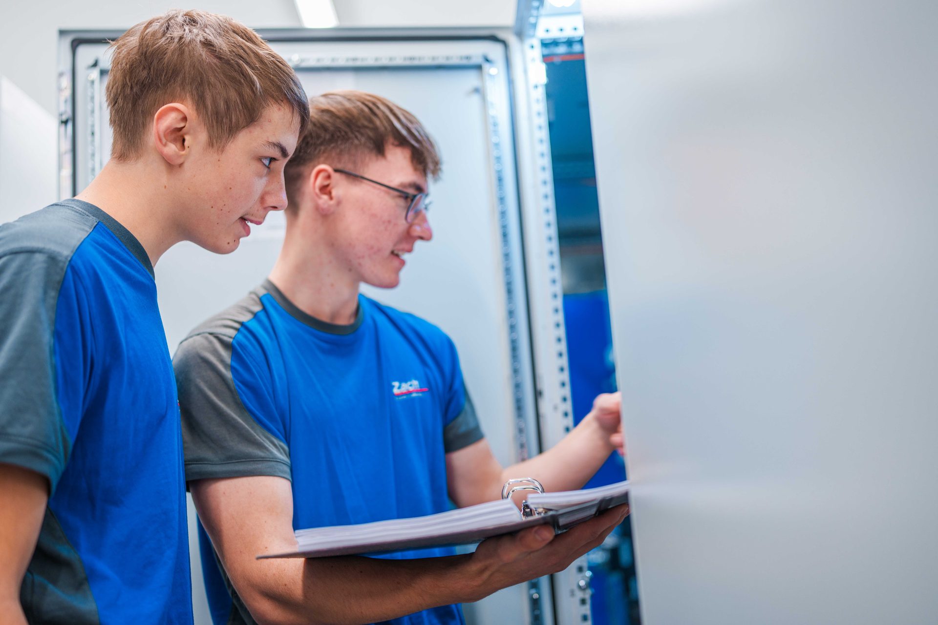 Two young men in blue shirts examine an open electrical cabinet, one holding a binder.