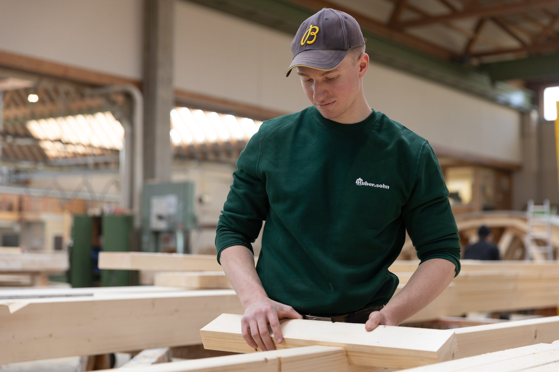 Young carpenter in a green sweatshirt and cap working with wooden beams in a workshop.