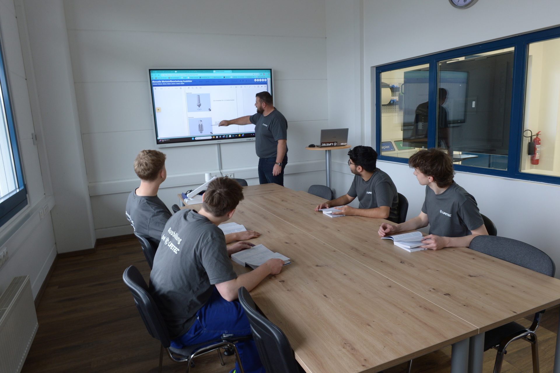 Man presenting on screen to four male students in a modern classroom.