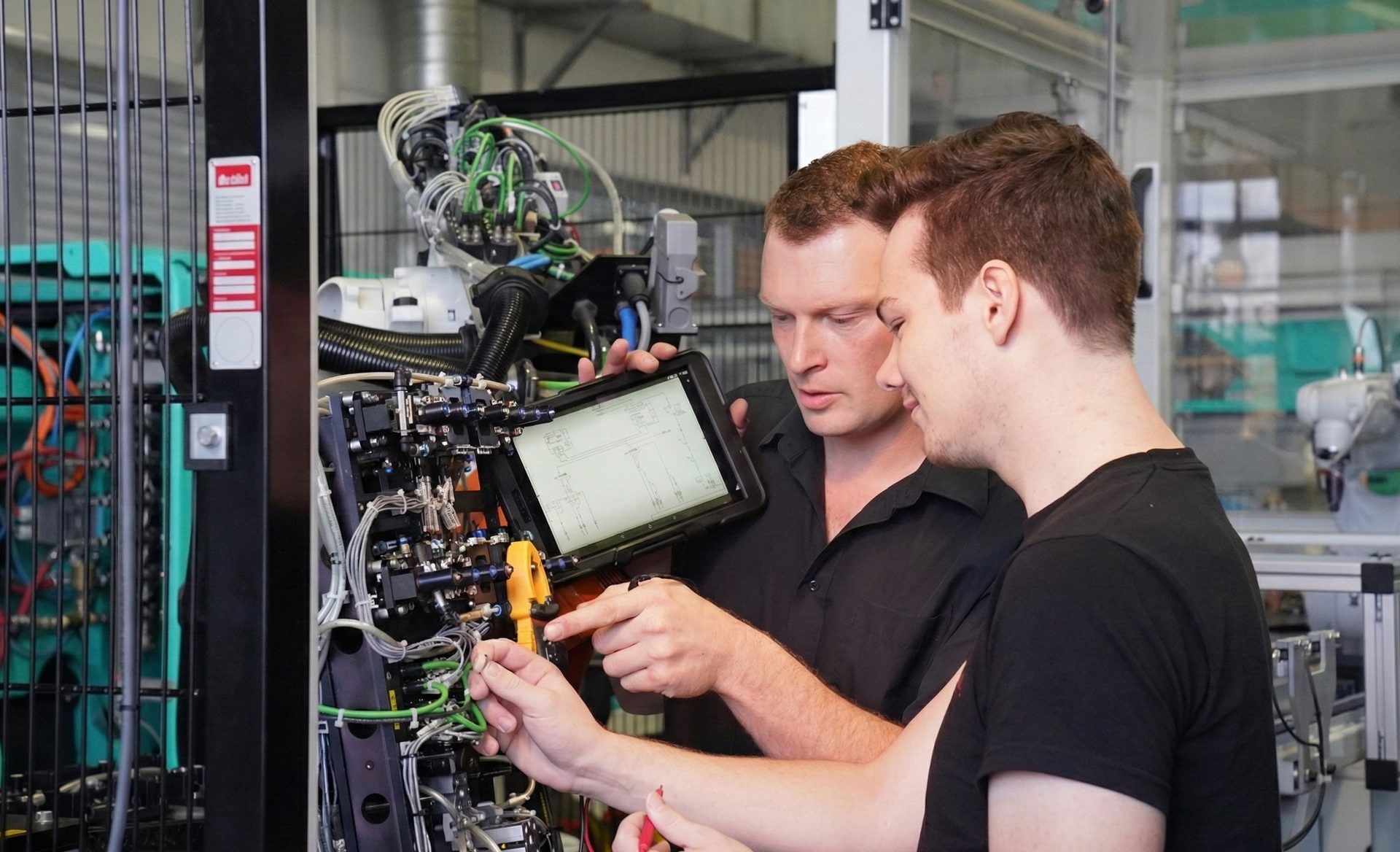 Two men collaborate on intricate machinery, using a tablet displaying schematics.