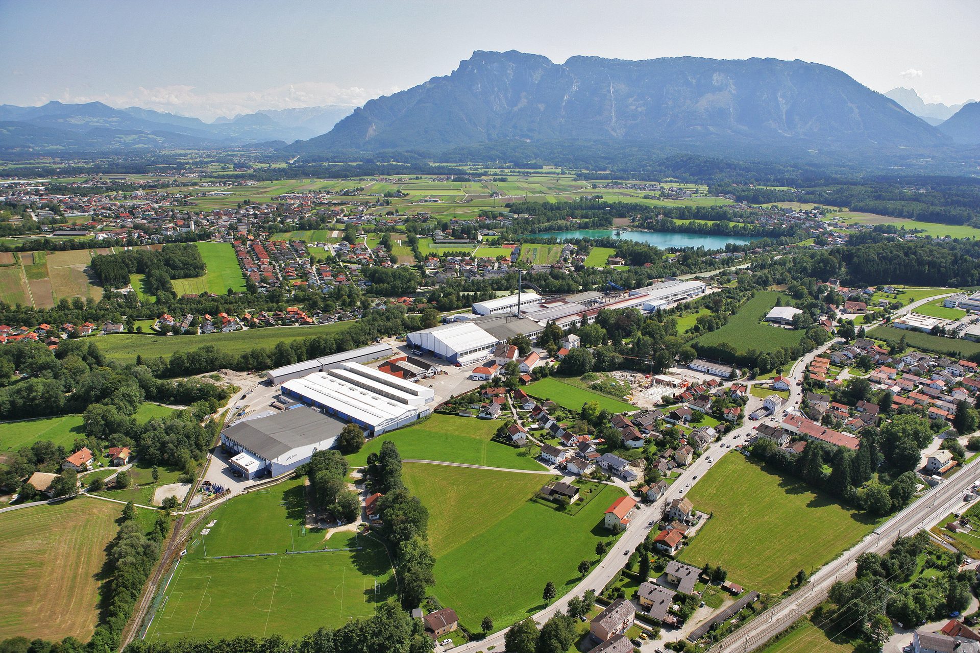 Aerial view: Alpine valley with town, factory buildings, lake, and mountains.