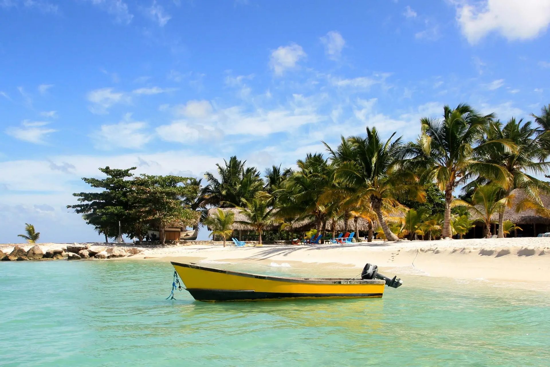 Yellow boat in clear turquoise water by a sandy beach with palm trees and blue sky.