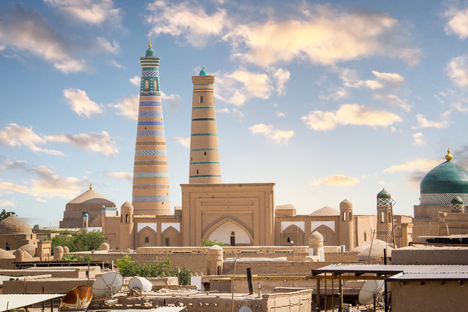Two decorated minarets rise above a historic city with domes under a cloudy sky.