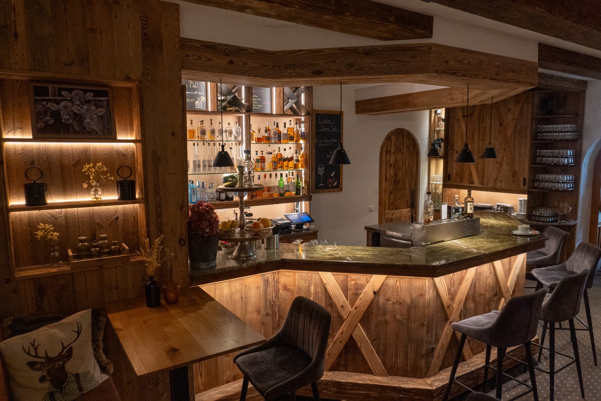 Cozy, rustic wooden bar and lounge area with illuminated shelves of bottles and seating.