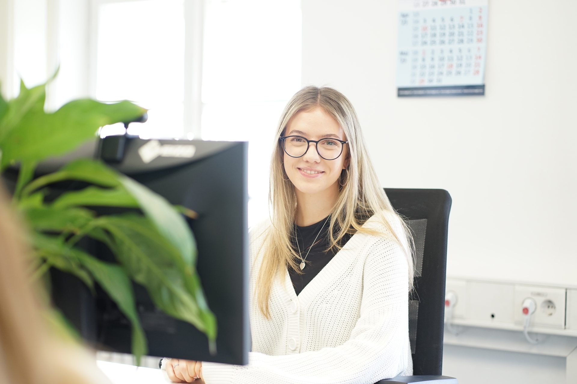 Smiling woman with glasses sits at a desk with a computer in an office.