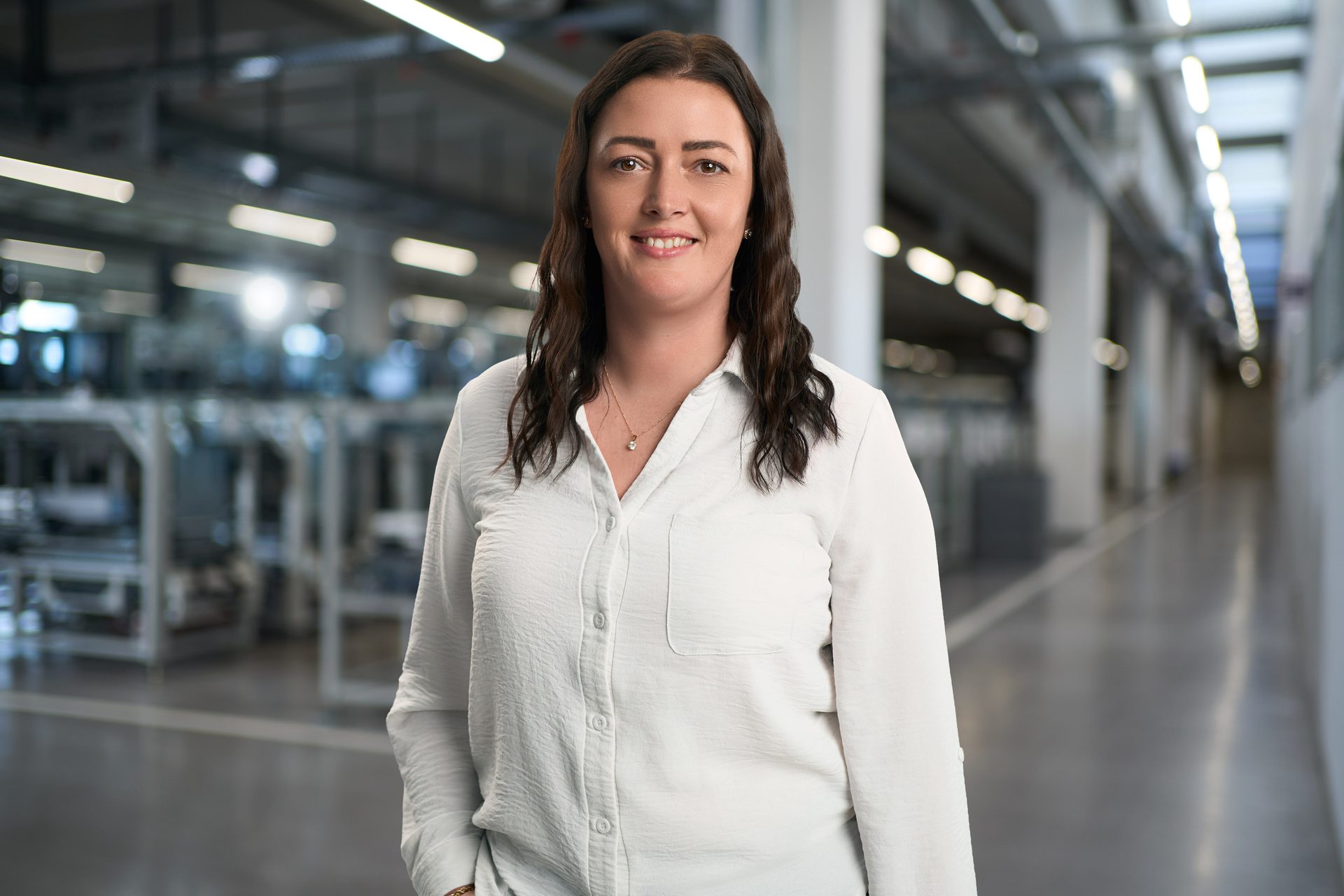 A smiling woman with dark hair in a white shirt stands in a modern factory setting, looking at the camera.