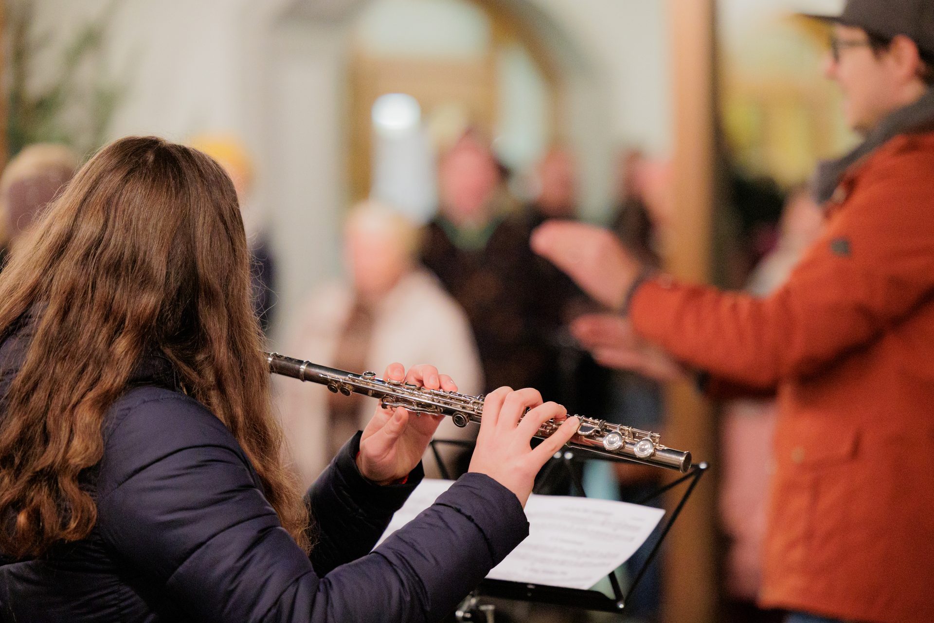 A person with long brown hair plays a flute, reading sheet music, with blurry people in the background.