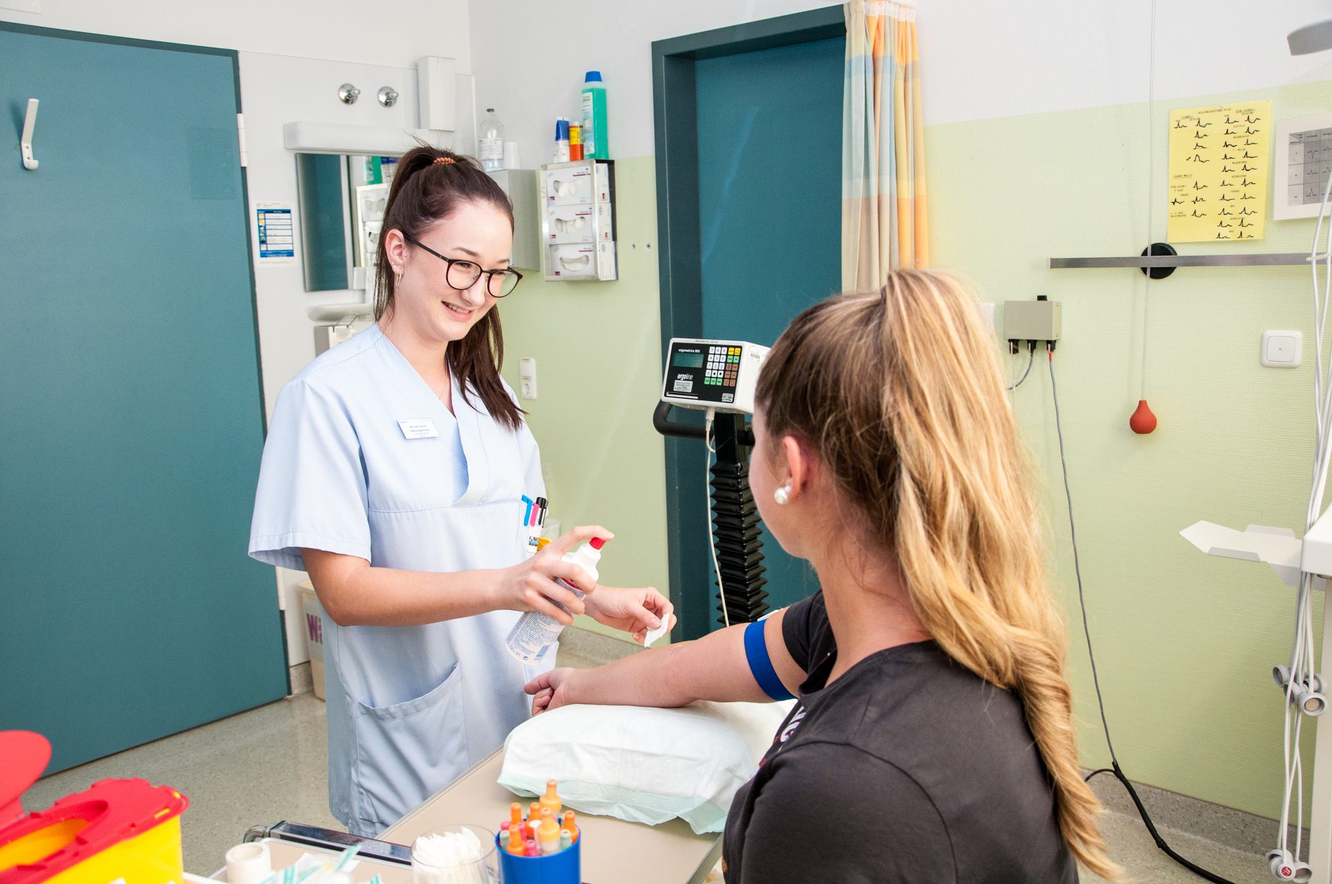 A smiling nurse disinfects a patient's arm, tourniquet in place, in a medical setting.