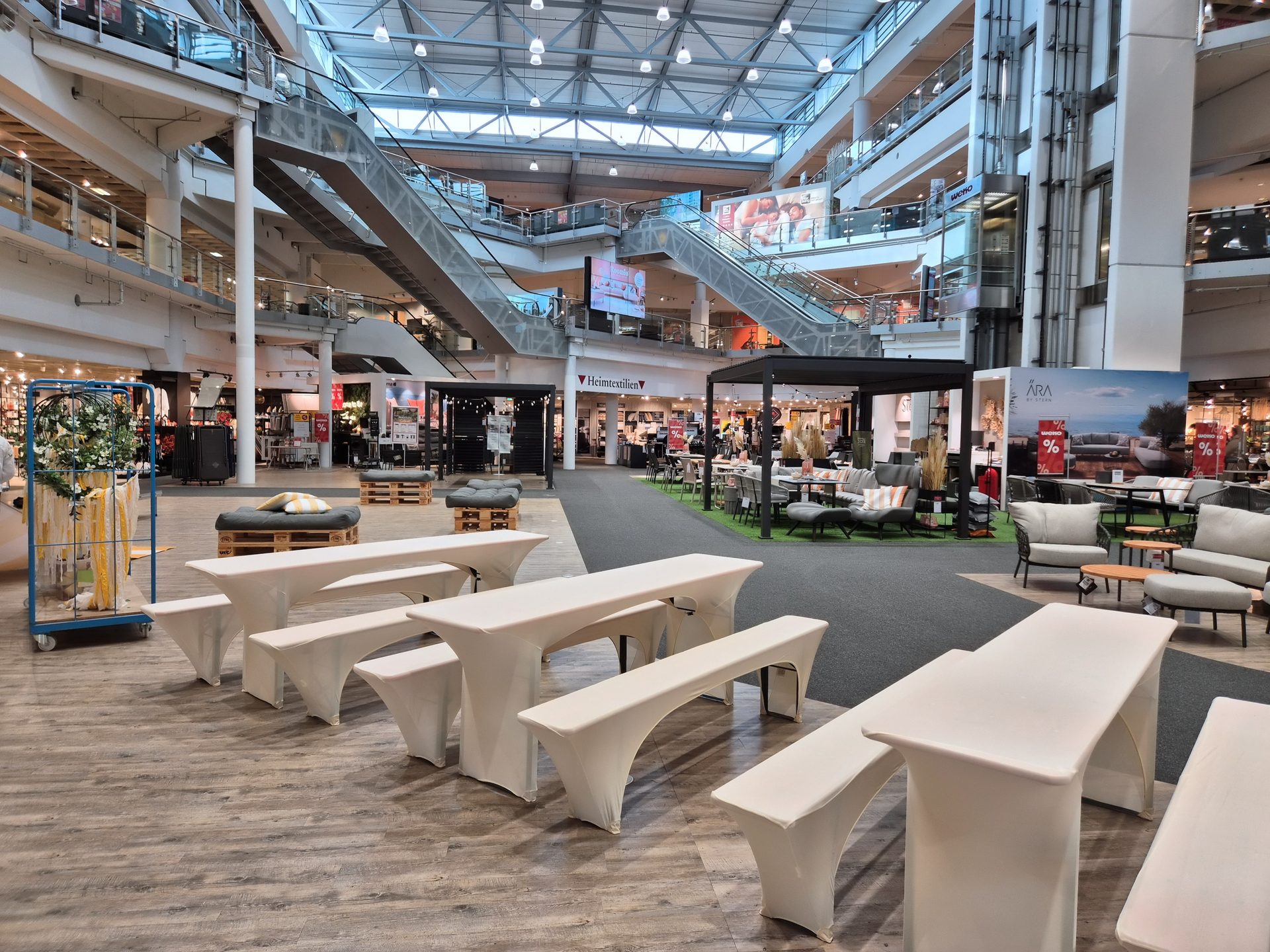A large, multi-story furniture store interior with escalators, various displays, and white event tables.