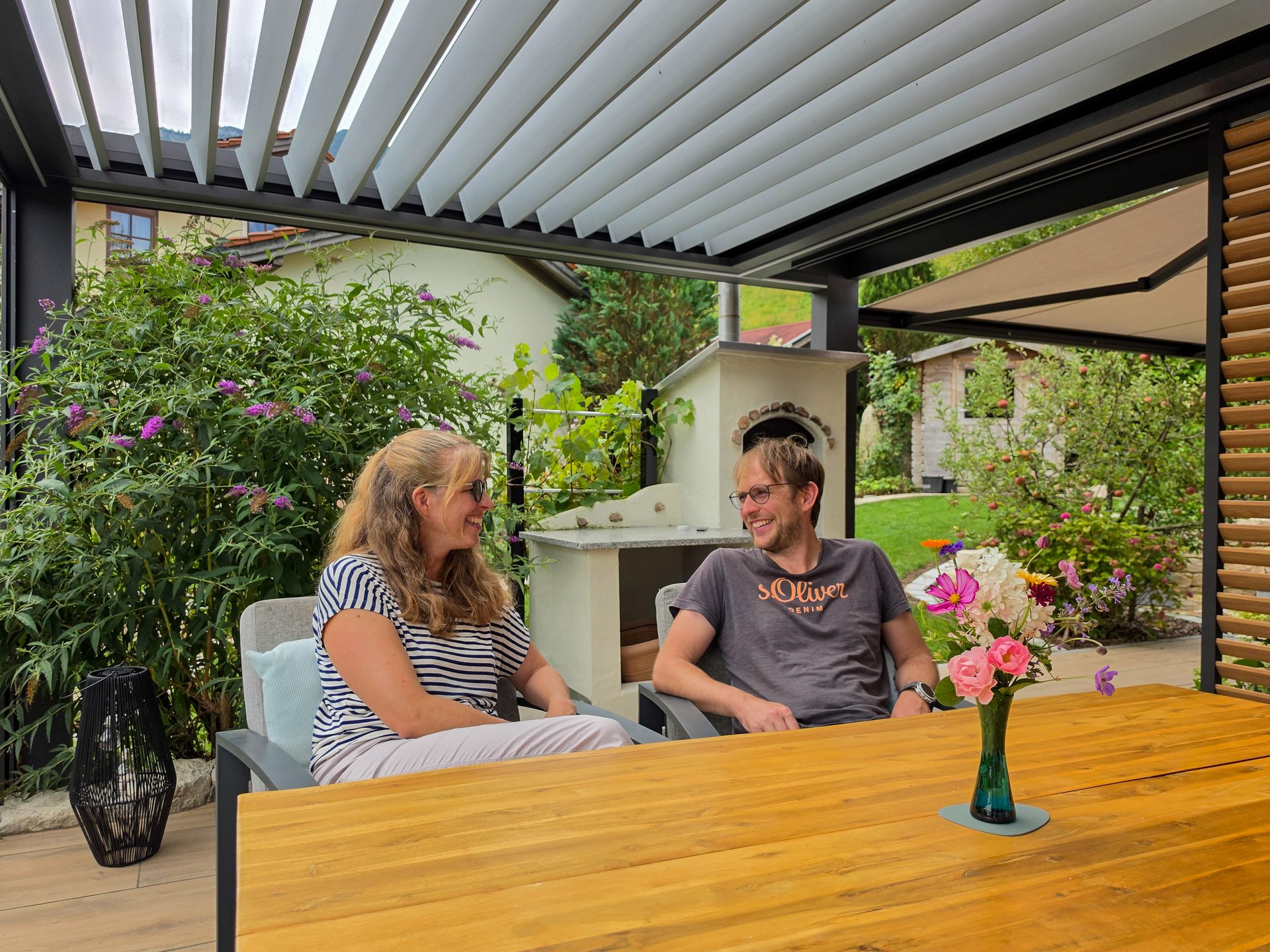 A smiling couple relaxing on a modern patio under a pergola, enjoying their lush garden.