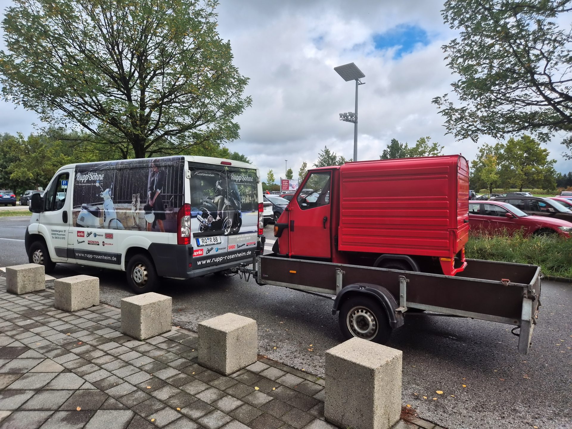 White van towing a red three-wheeled utility vehicle on a trailer in a parking lot.