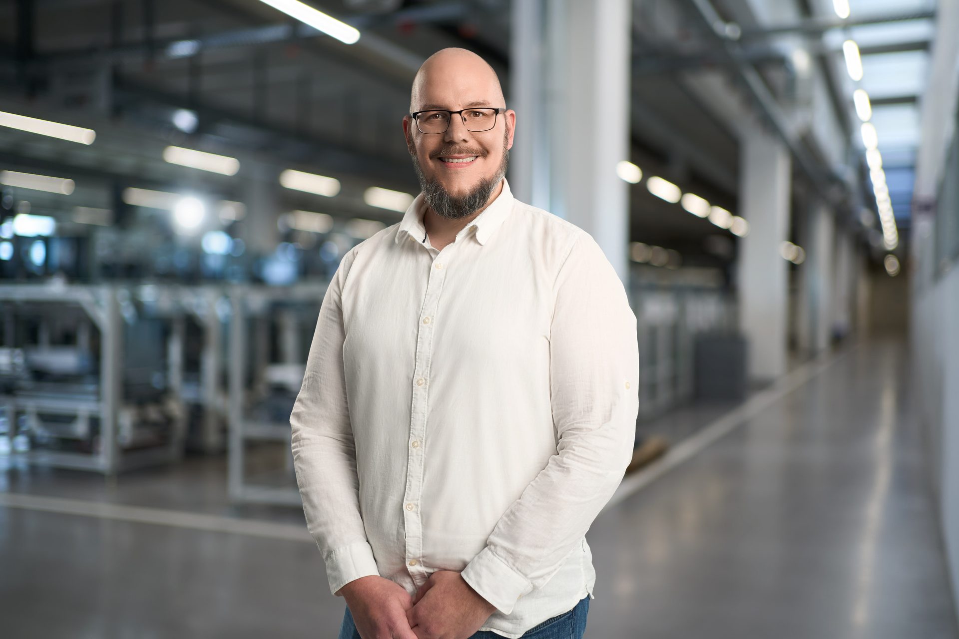 A bald man with a beard and glasses smiles, wearing a white shirt in a modern factory setting.