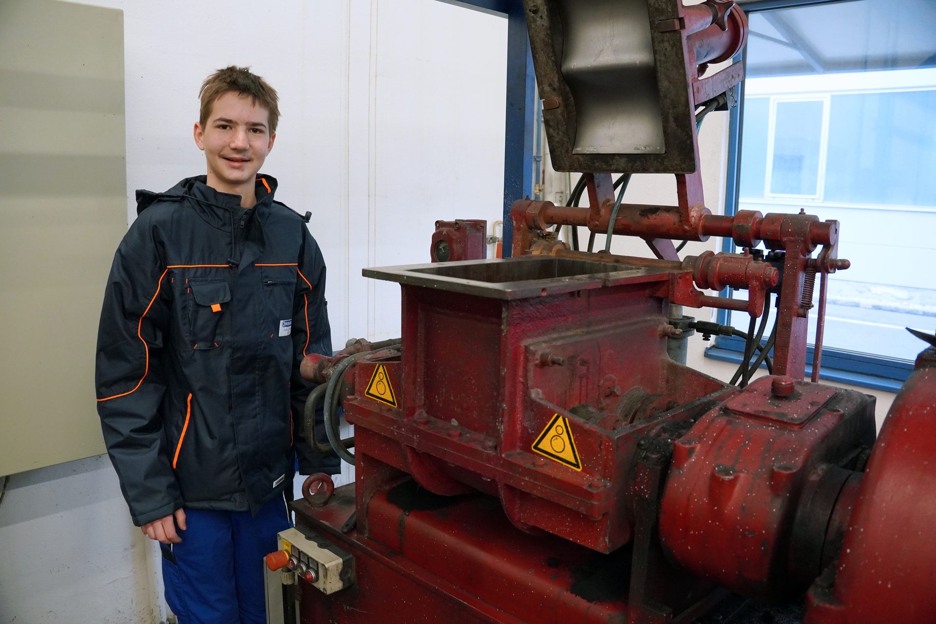 A young man in a dark work uniform stands beside a large red industrial machine.