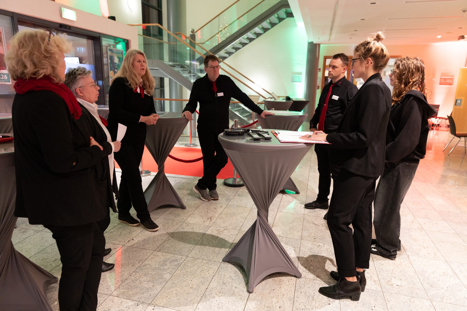 People (some staff) around cocktail tables in a modern event lobby with a staircase and colorful lighting.