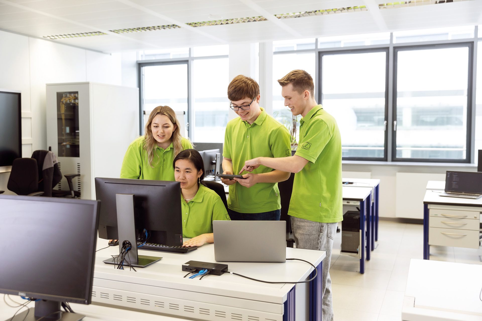 Four students in green shirts collaborating on computers and a tablet in an office.
