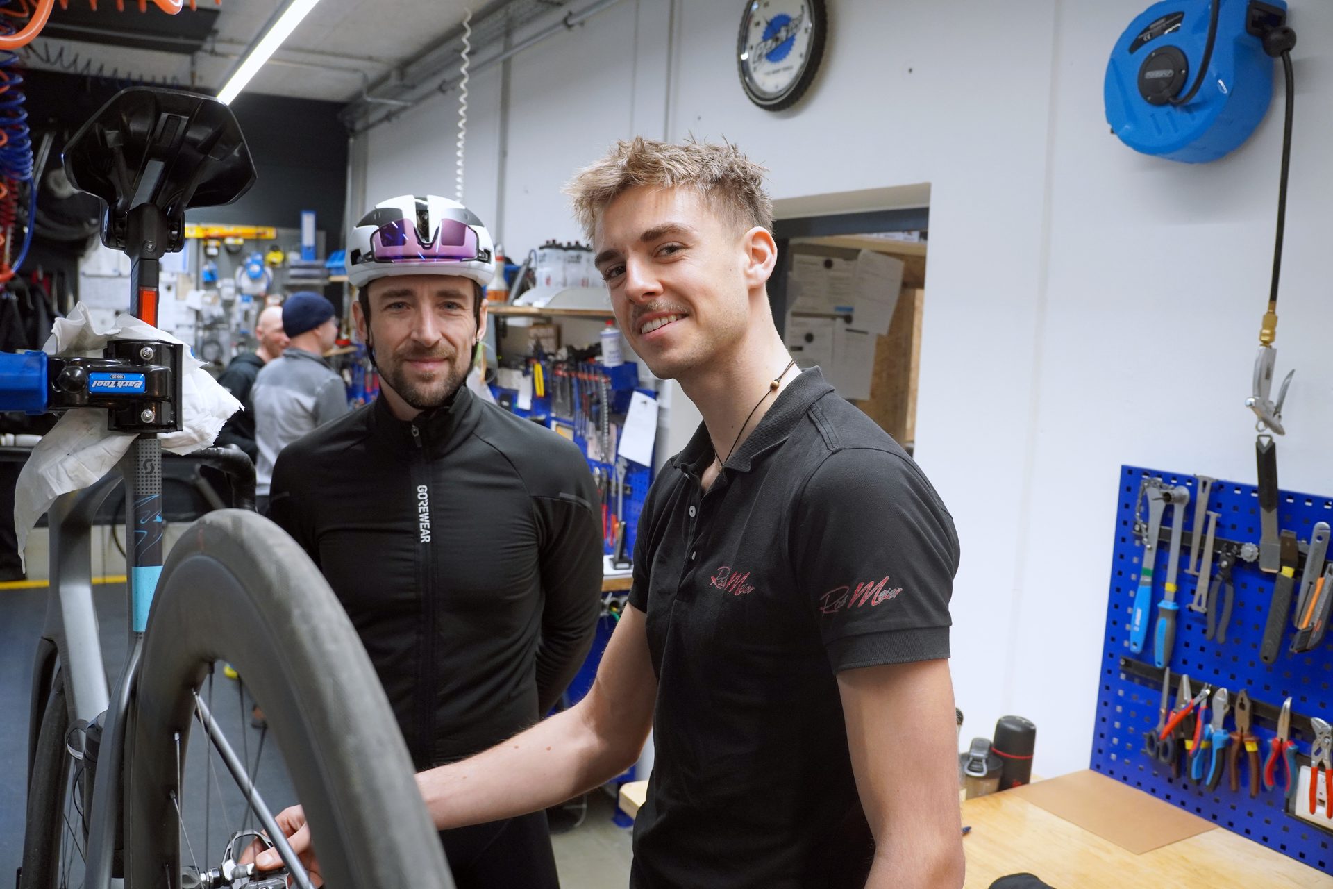 Two men, one in a cycling helmet, the other in a polo shirt, smiling in a bike workshop.