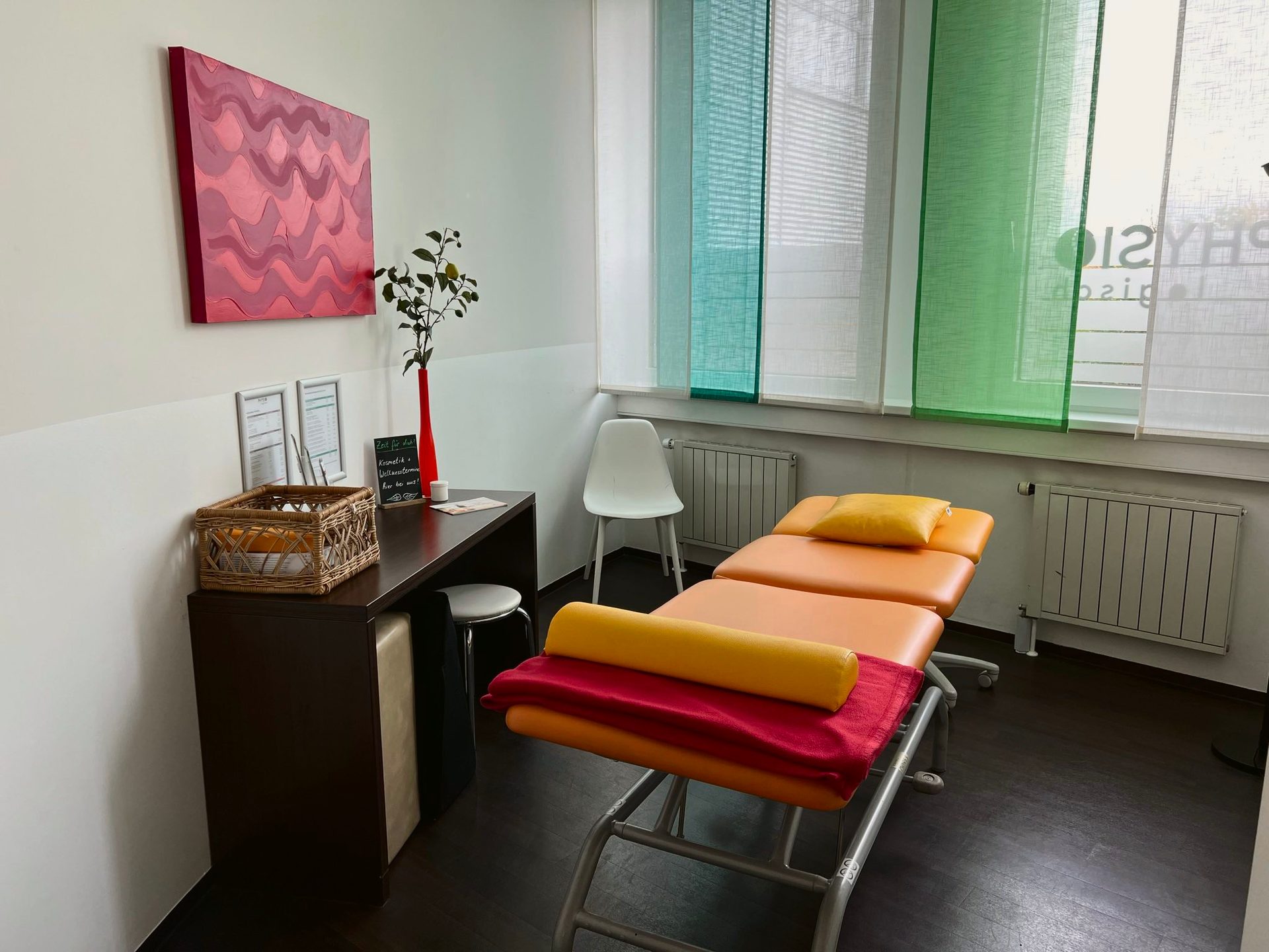 Physiotherapy room with an orange treatment bed, a dark wood desk, and colorful window blinds.