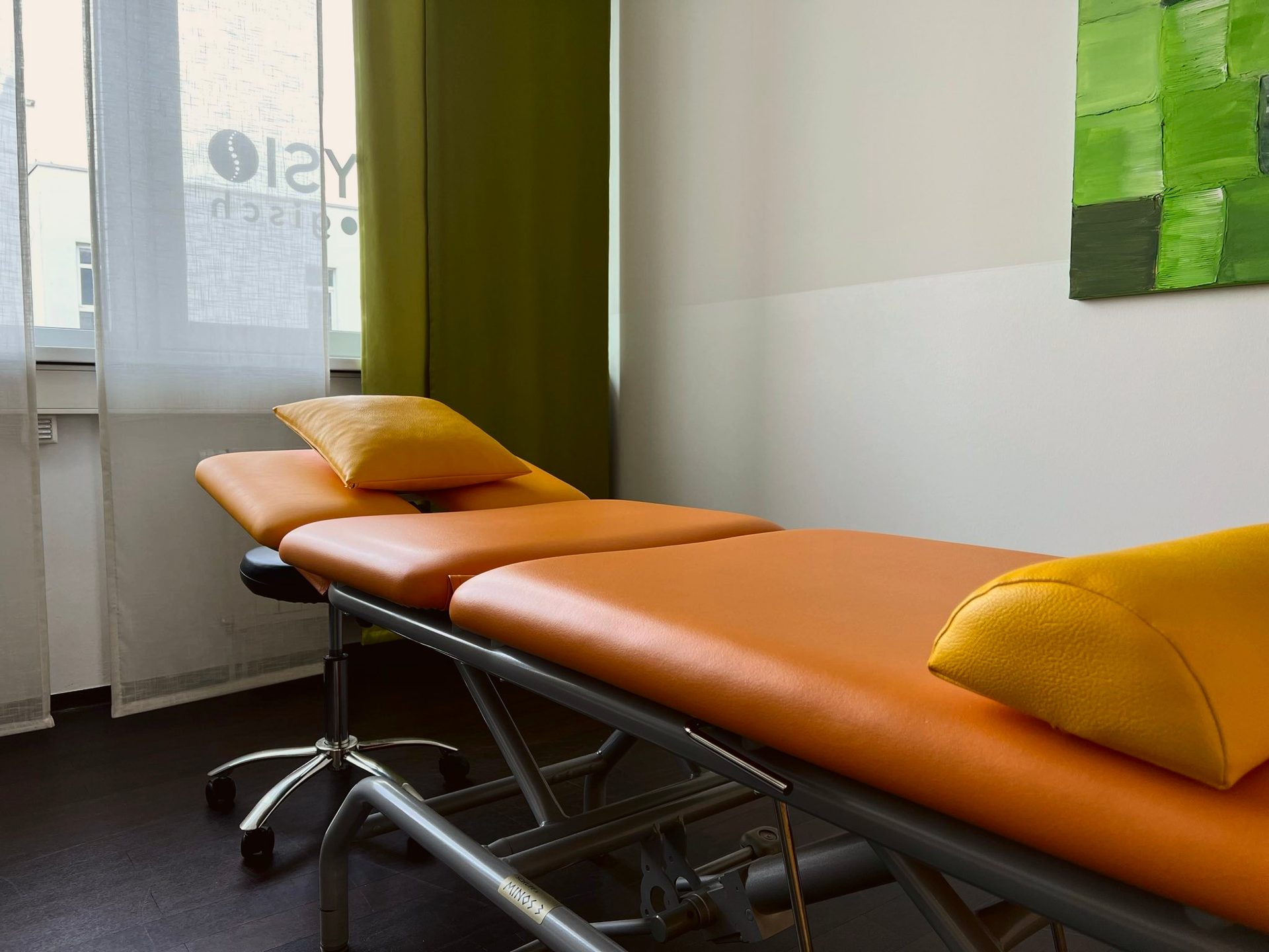 An empty physical therapy room with an orange examination table, yellow pillows, and green and white walls.