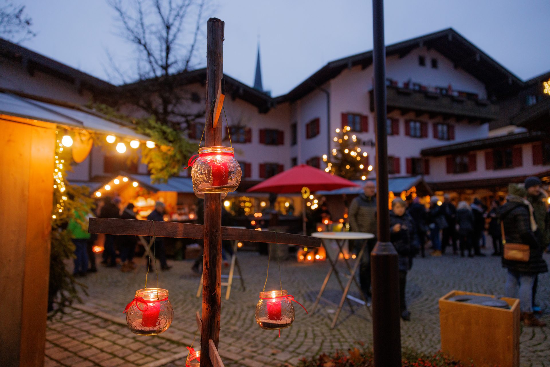 A festive Christmas market scene at dusk with hanging red candle lanterns, people, and illuminated buildings.