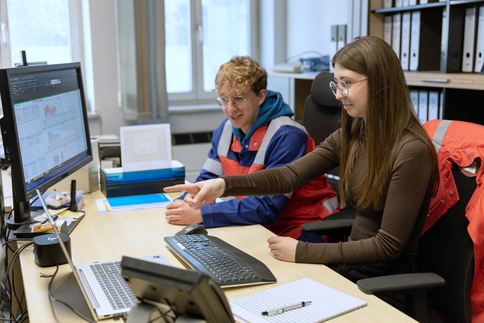 Two people look at a computer screen with data and charts in an office.