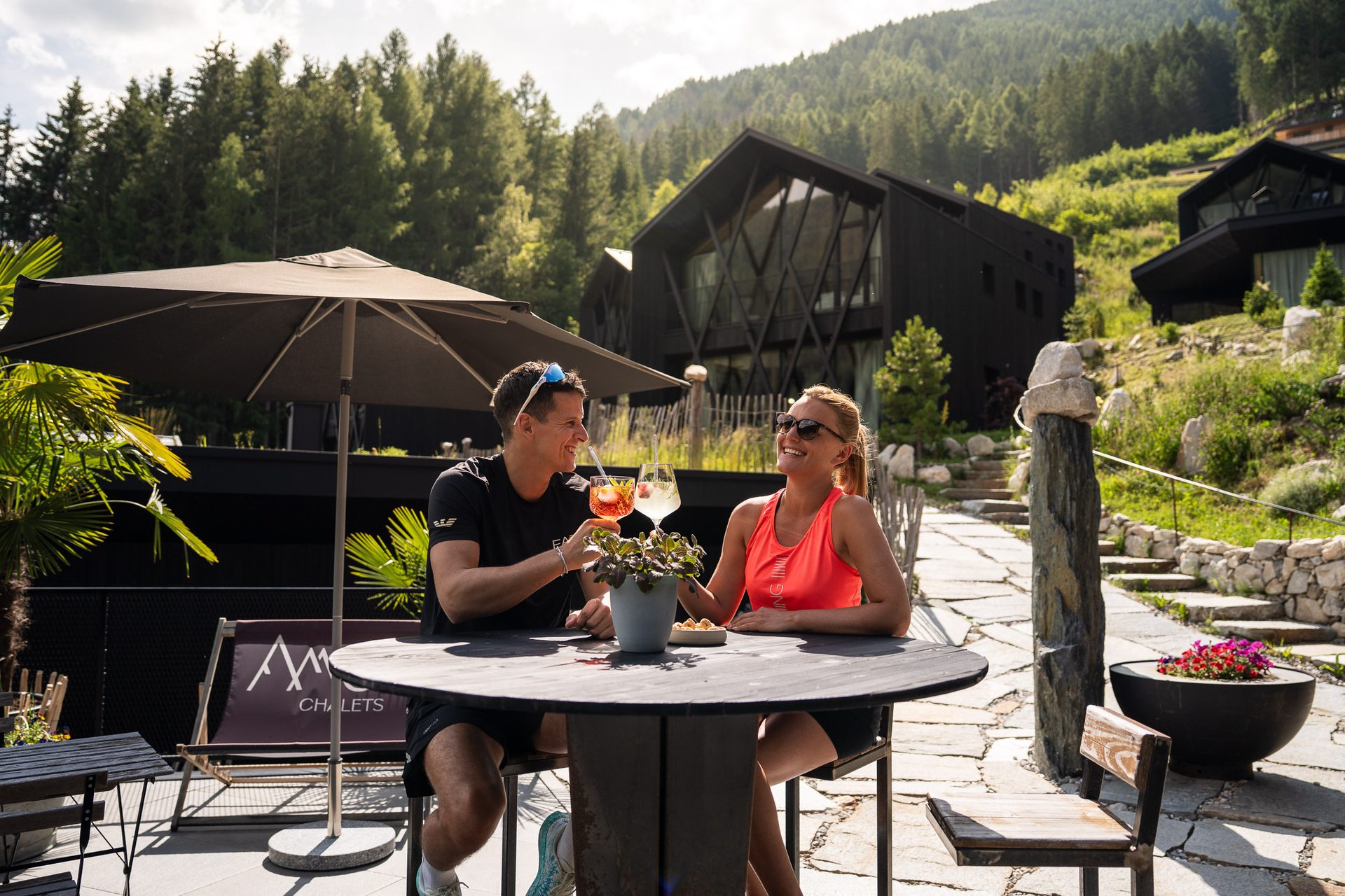 A couple cheers with cocktails at an outdoor table in a mountain resort with modern black chalets.