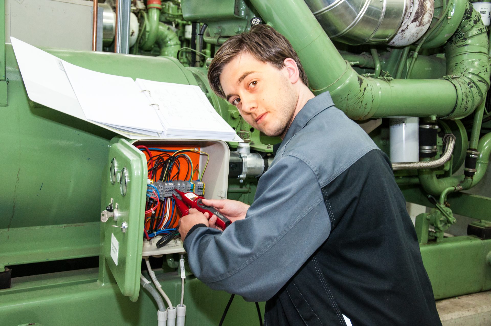 Technician troubleshooting an electrical panel with a multimeter, looking at the viewer.