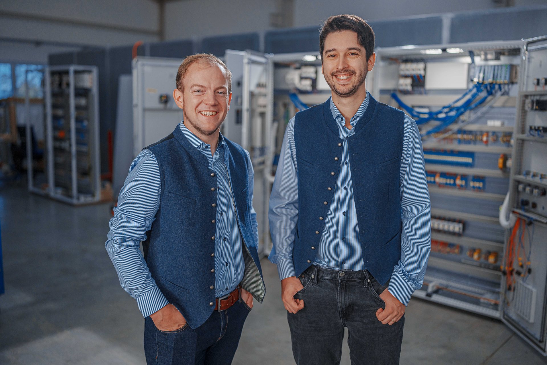 Two smiling men in blue vests and striped shirts stand in an industrial electrical workshop.