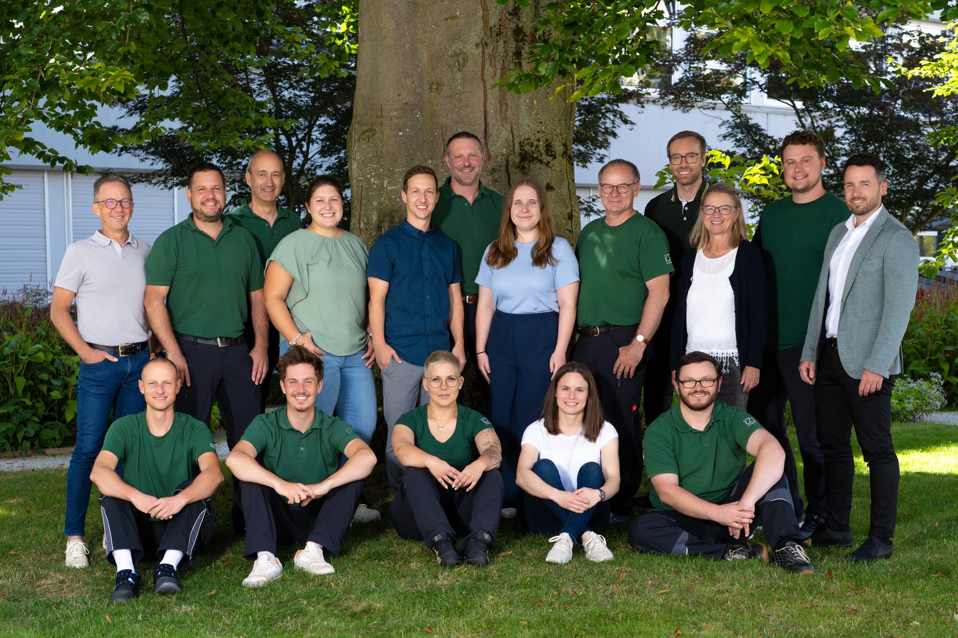 A group of 17 people, men and women of various ages, smiling outdoors on grass in front of a large tree.