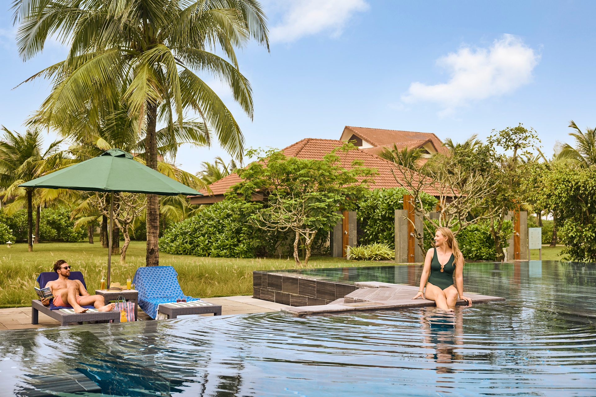 A man reads on a lounge chair and a woman sits in a pool at a tropical resort.