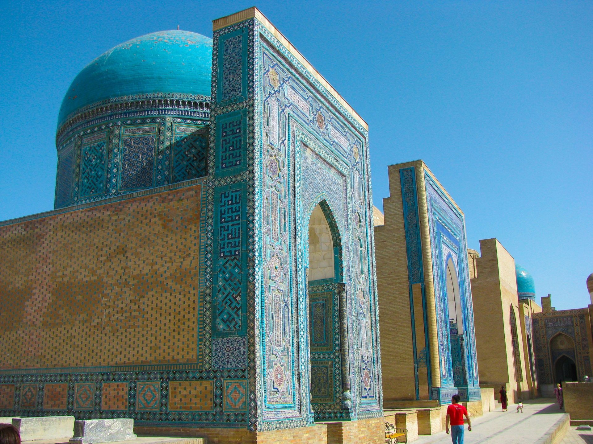 Intricate blue-tiled Islamic architecture with a large dome under a clear sky. A person walks by.