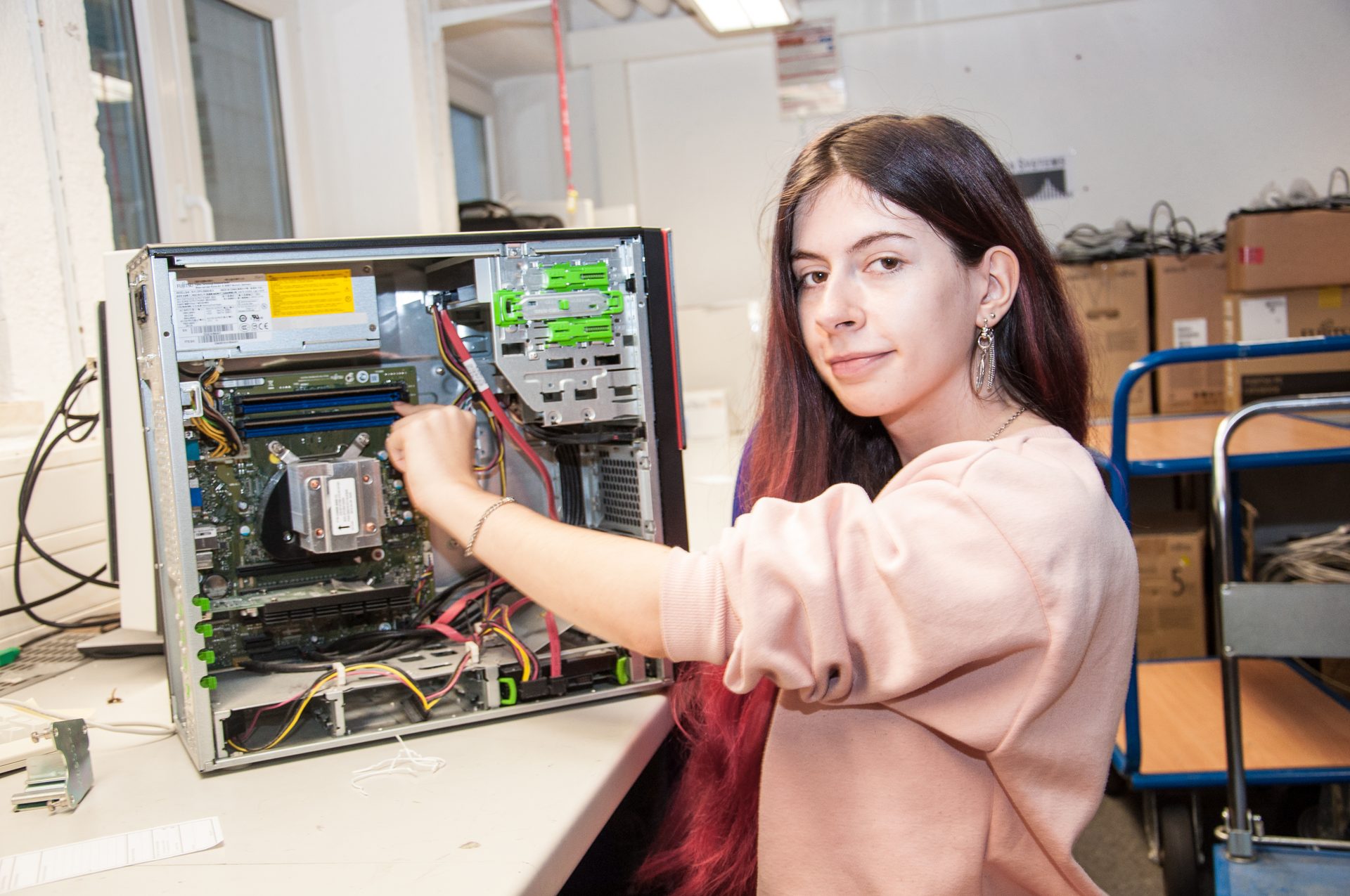 A young woman works on the internal components of a desktop computer, looking at the viewer.