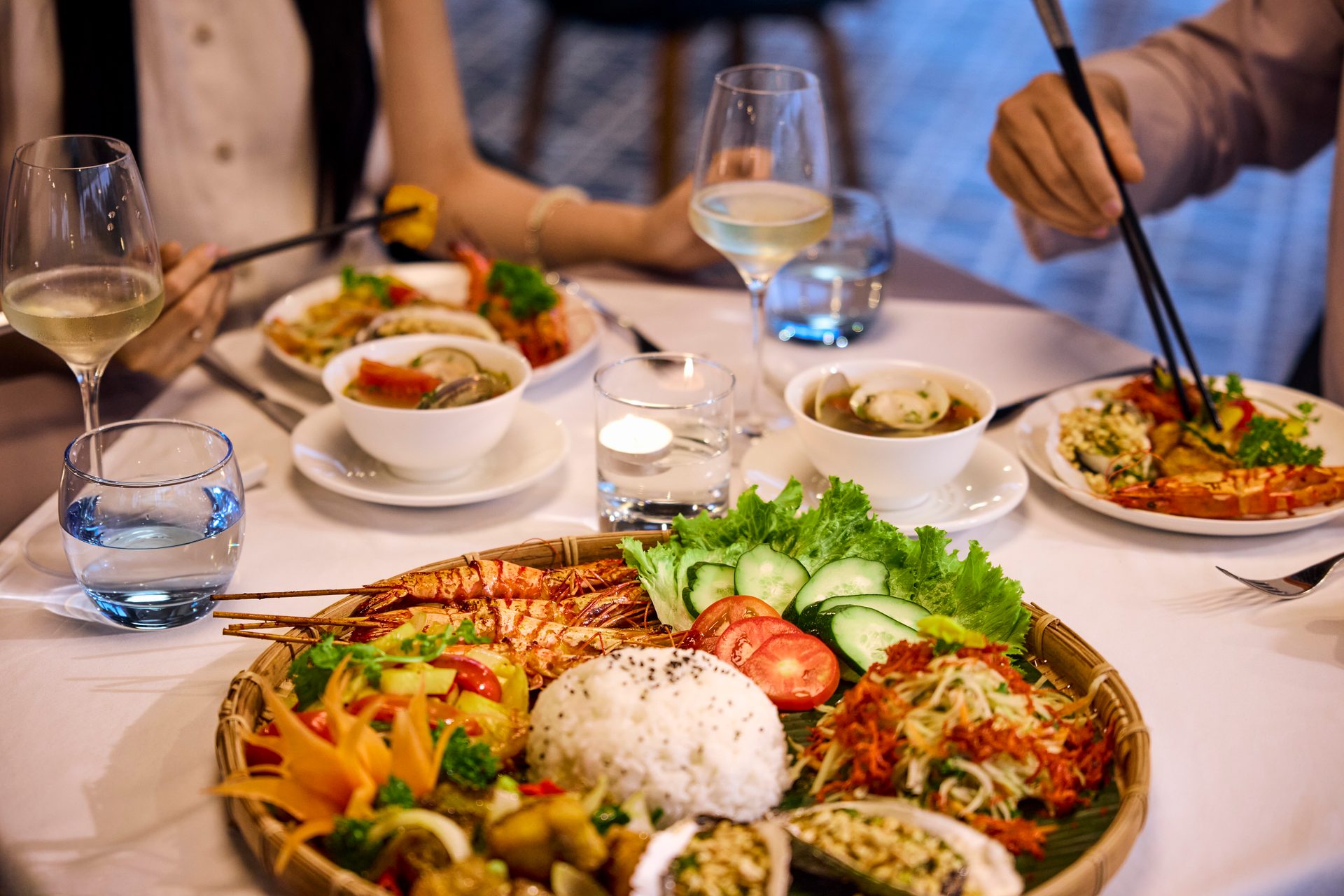 Two diners enjoying a Vietnamese meal with grilled shrimp, rice, salads, soups, and wine on a table.