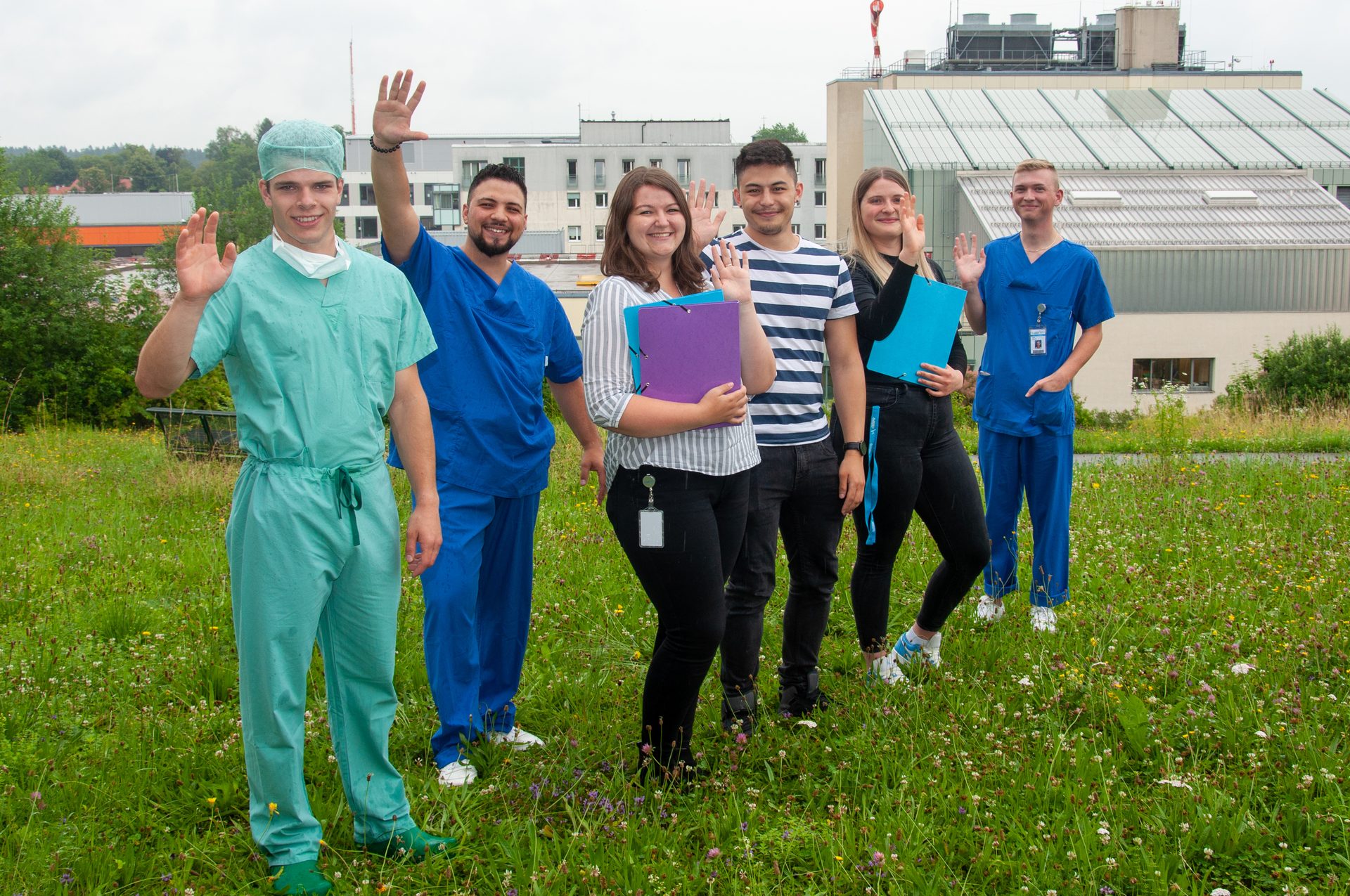 Six people, some in medical scrubs, wave from a grassy field with buildings in the background.