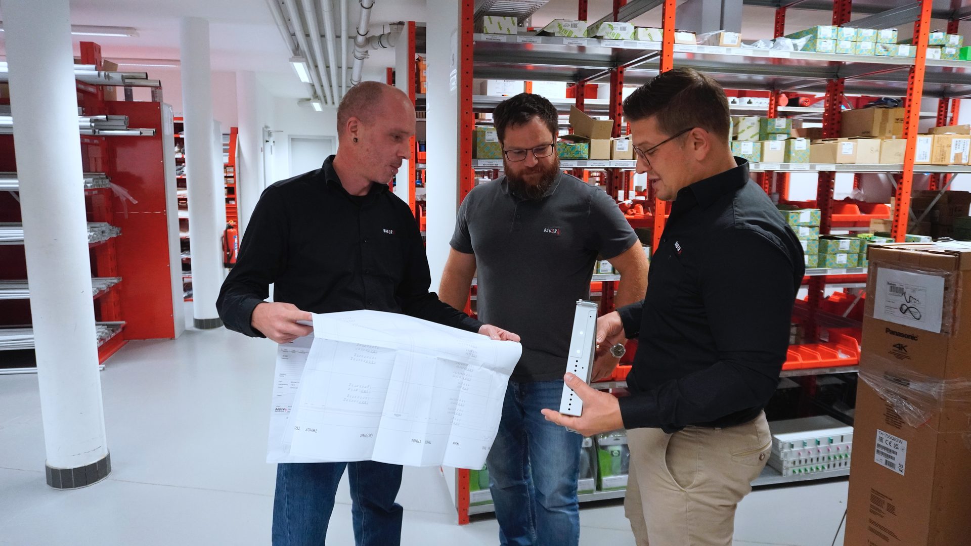 Three men in a warehouse examine a document and equipment, surrounded by storage shelves.