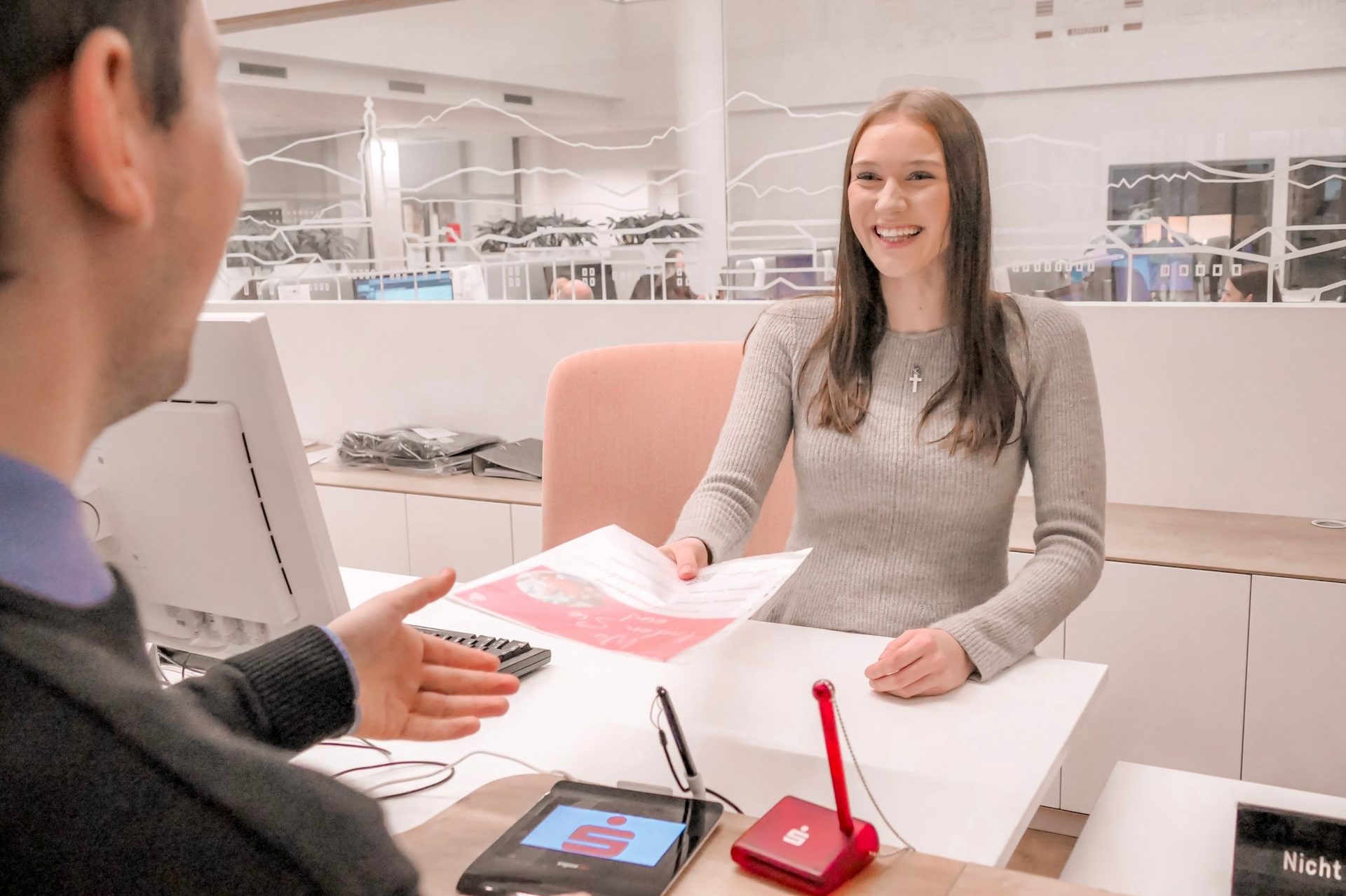 Smiling woman hands papers to a man across a desk in a modern office with Sparkasse branding.