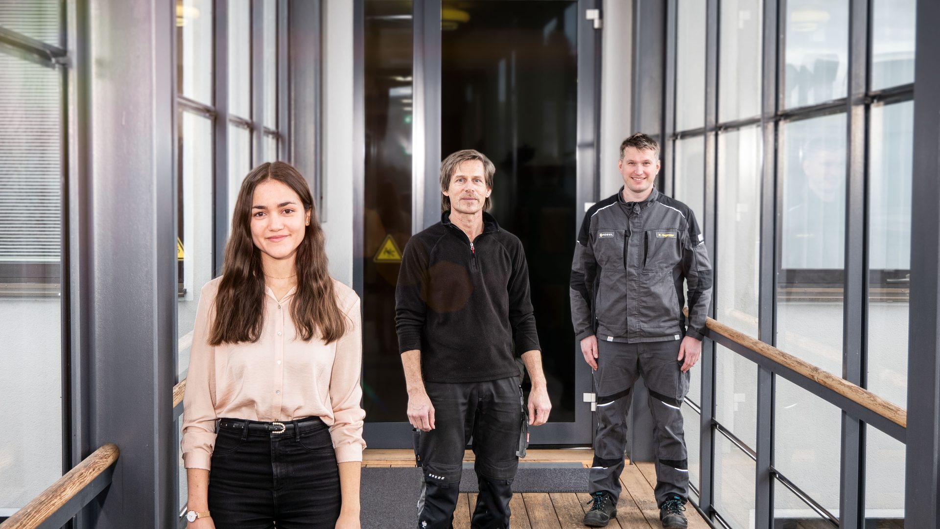 Three colleagues, a woman and two men, stand in a modern office hallway, smiling.