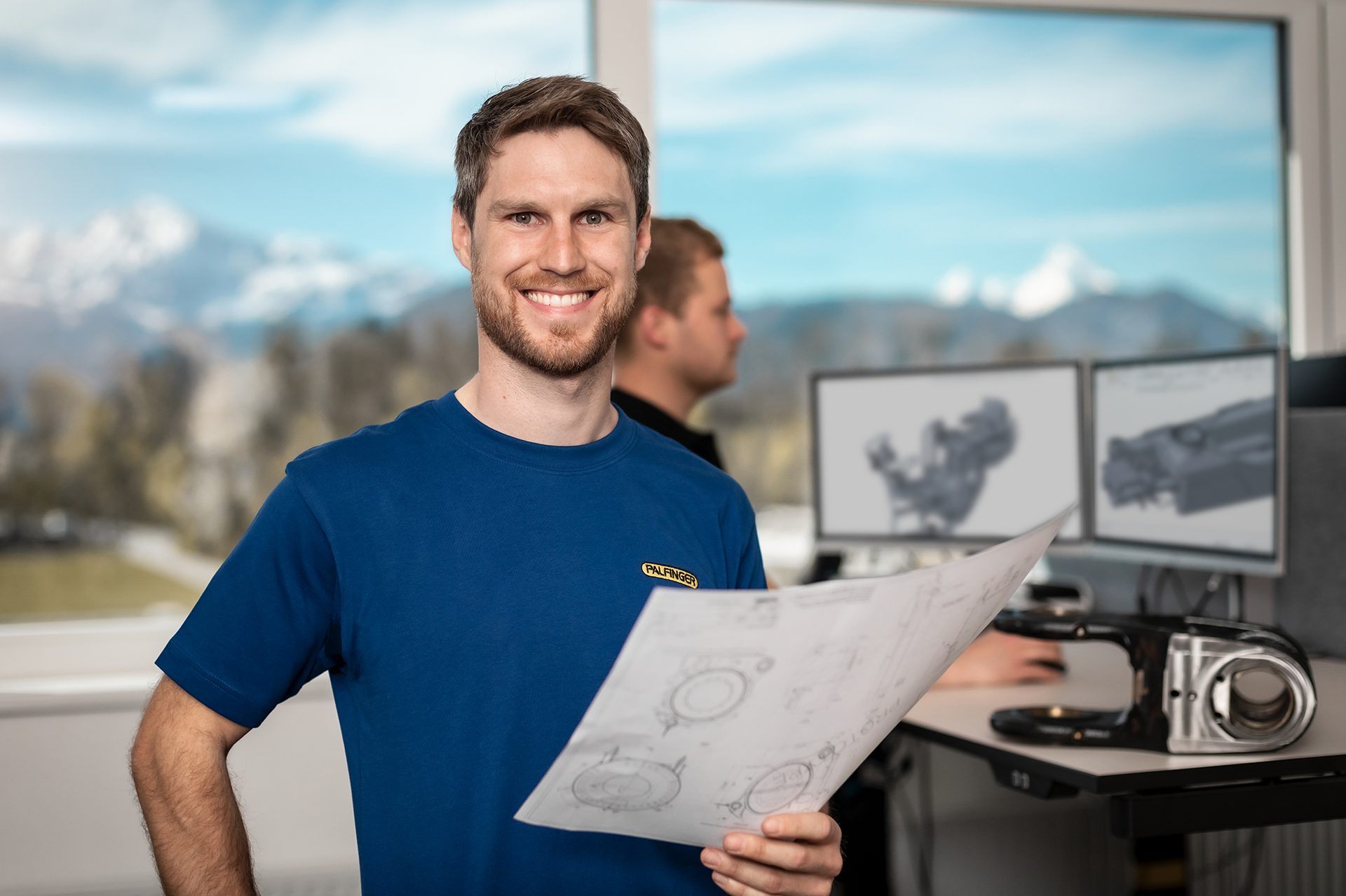 Smiling man in blue shirt holds blueprints, modern office with mountain view and CAD screens.