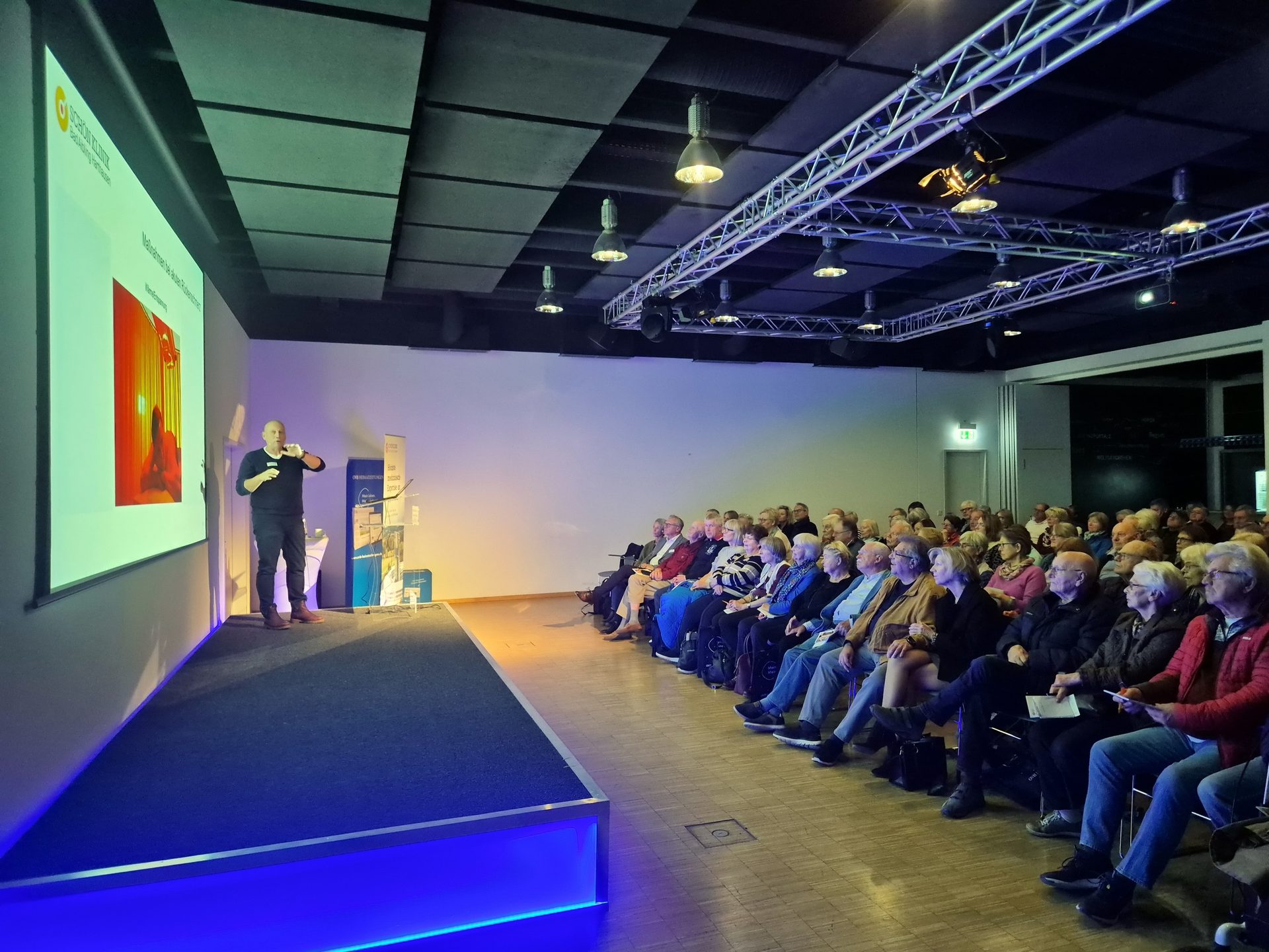 A speaker presents on a stage to an attentive audience in a large, dimly lit hall with stage lighting.