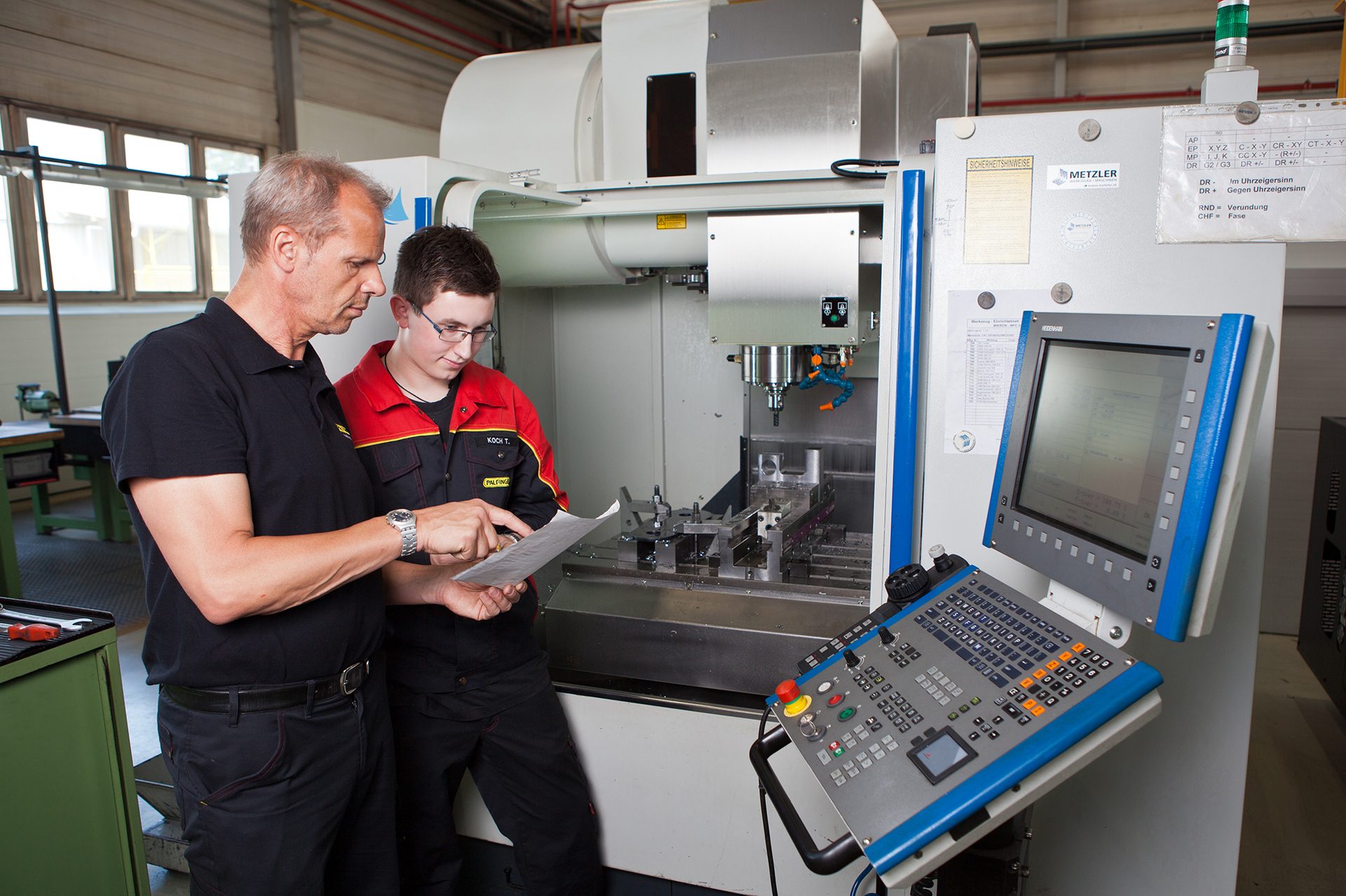 Two men in a workshop, one instructing the other at a CNC machine.