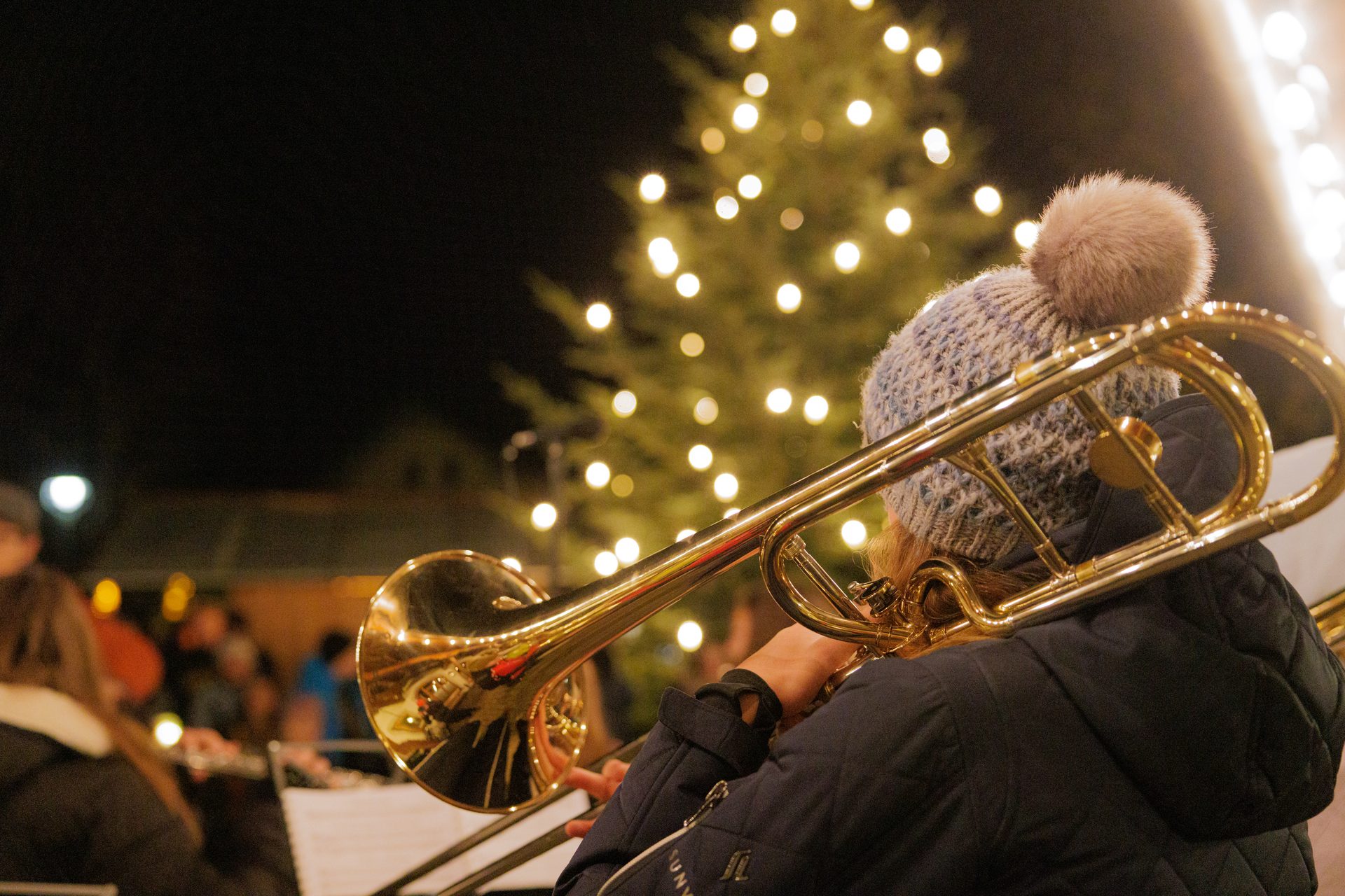 Person playing trombone at night, with a blurred Christmas tree and lights in the background.