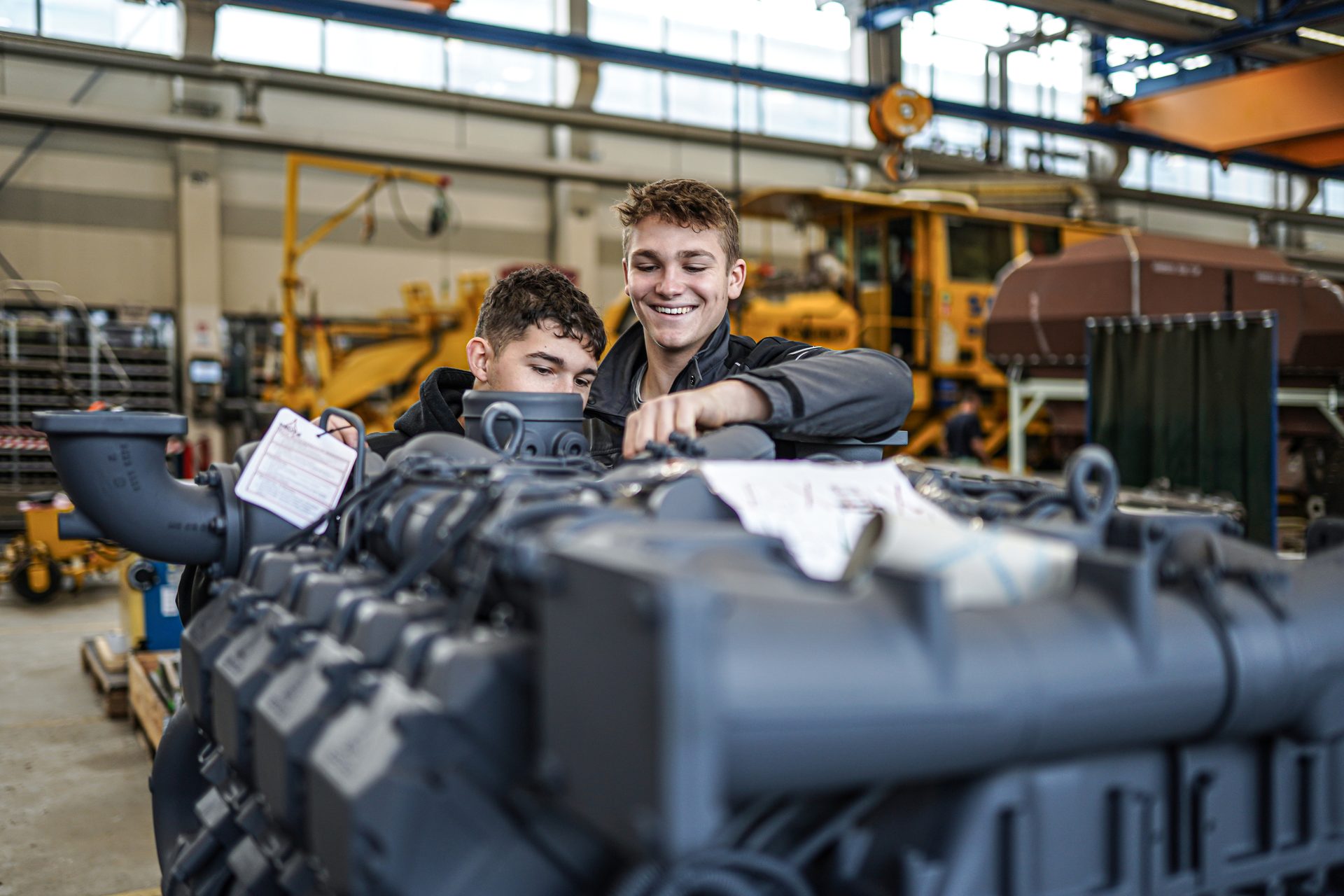 Two young men, possibly apprentices, inspect a large industrial engine in a workshop, smiling and engaged.