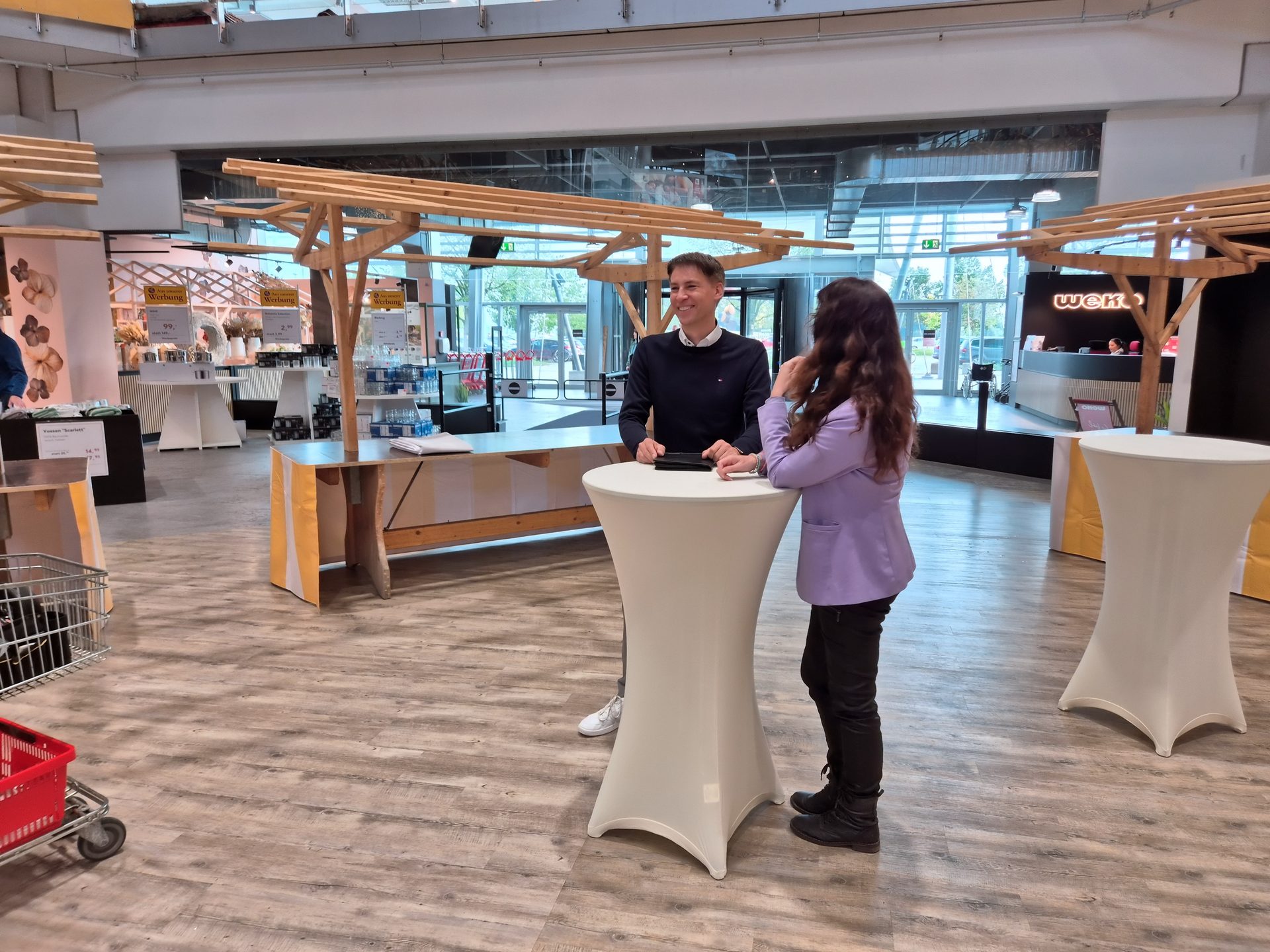 Two people talk at a high-top table in a modern store or exhibition space with wooden stalls.