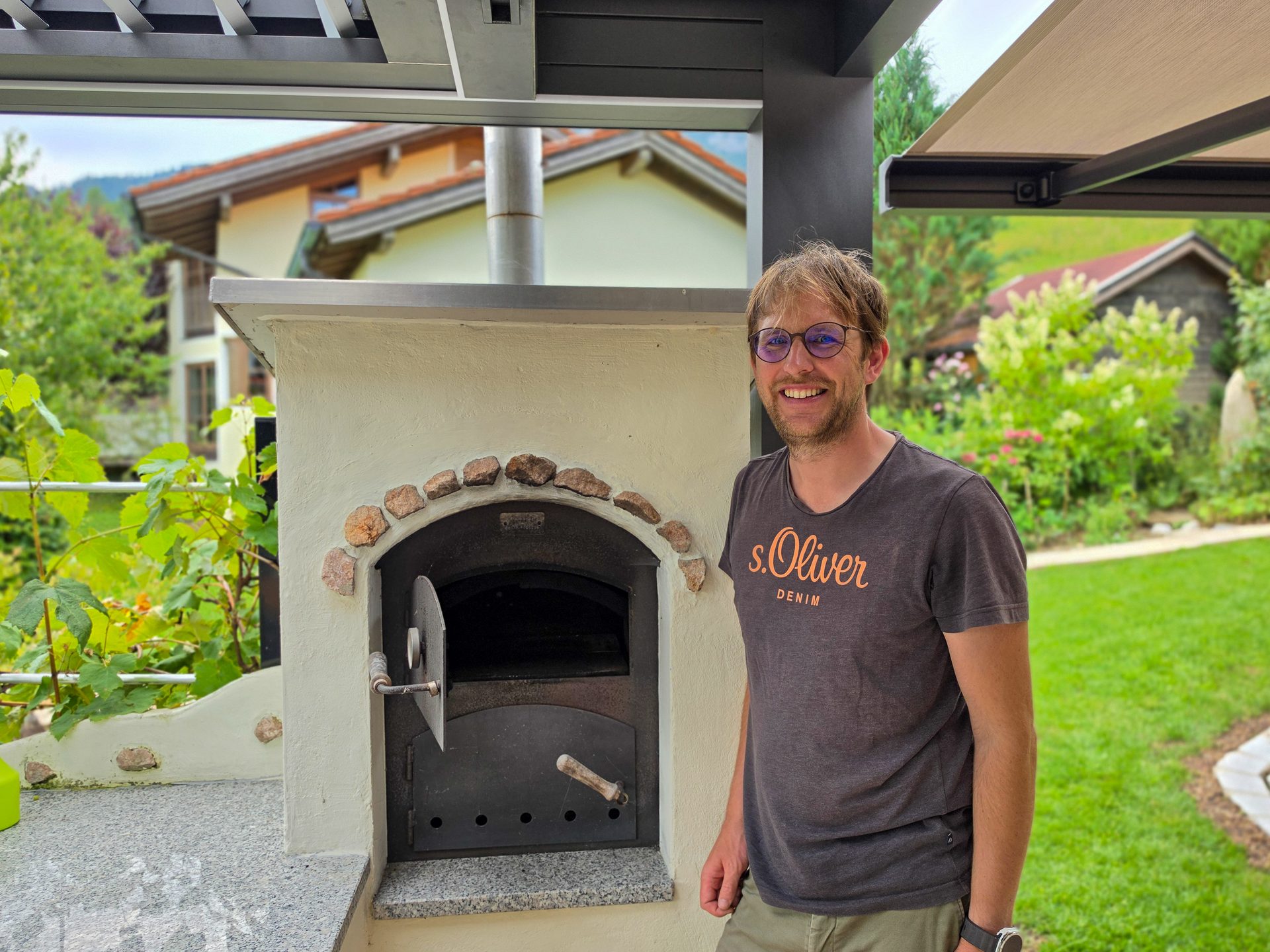 Smiling man standing next to an outdoor pizza oven in a sunny garden with green foliage and houses.
