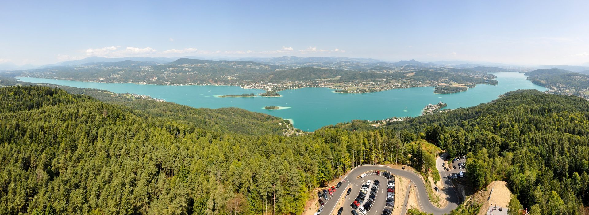 Aerial panorama of turquoise lake, green forests, distant mountains, and a winding road with parking.