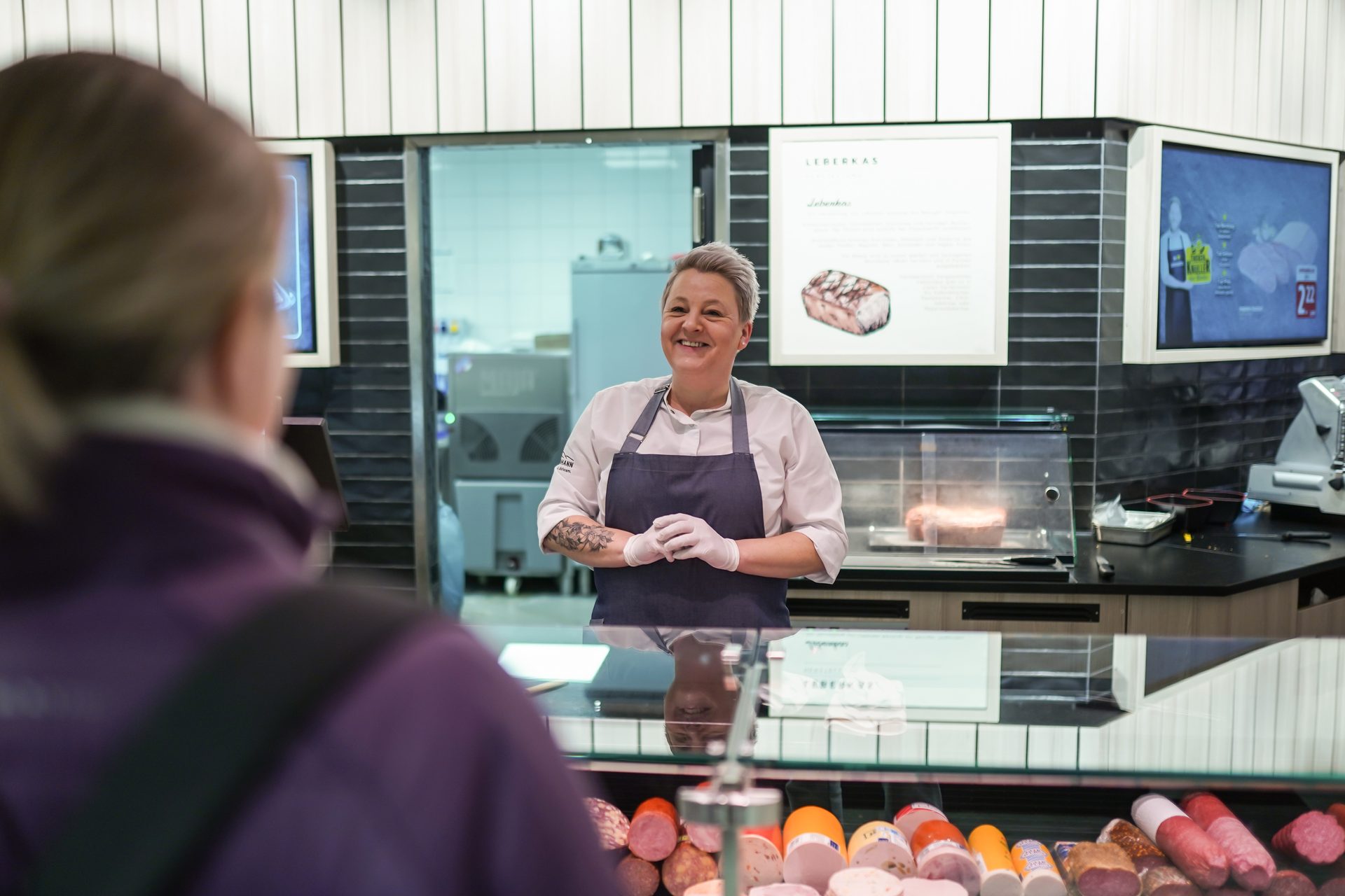 A smiling deli worker serves a customer at a counter displaying various meats.