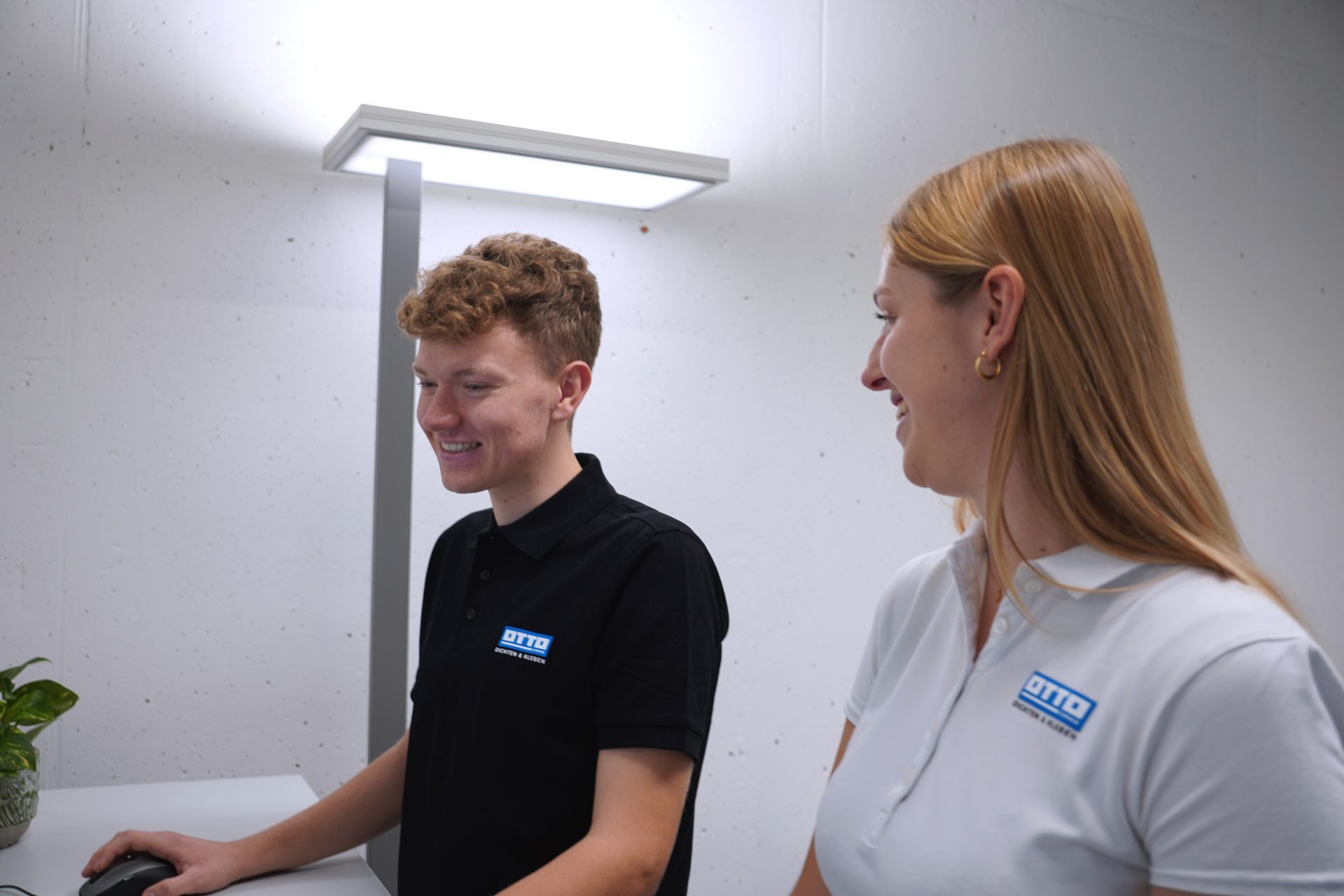 Smiling man and woman in OTTO polo shirts, man using mouse in office.