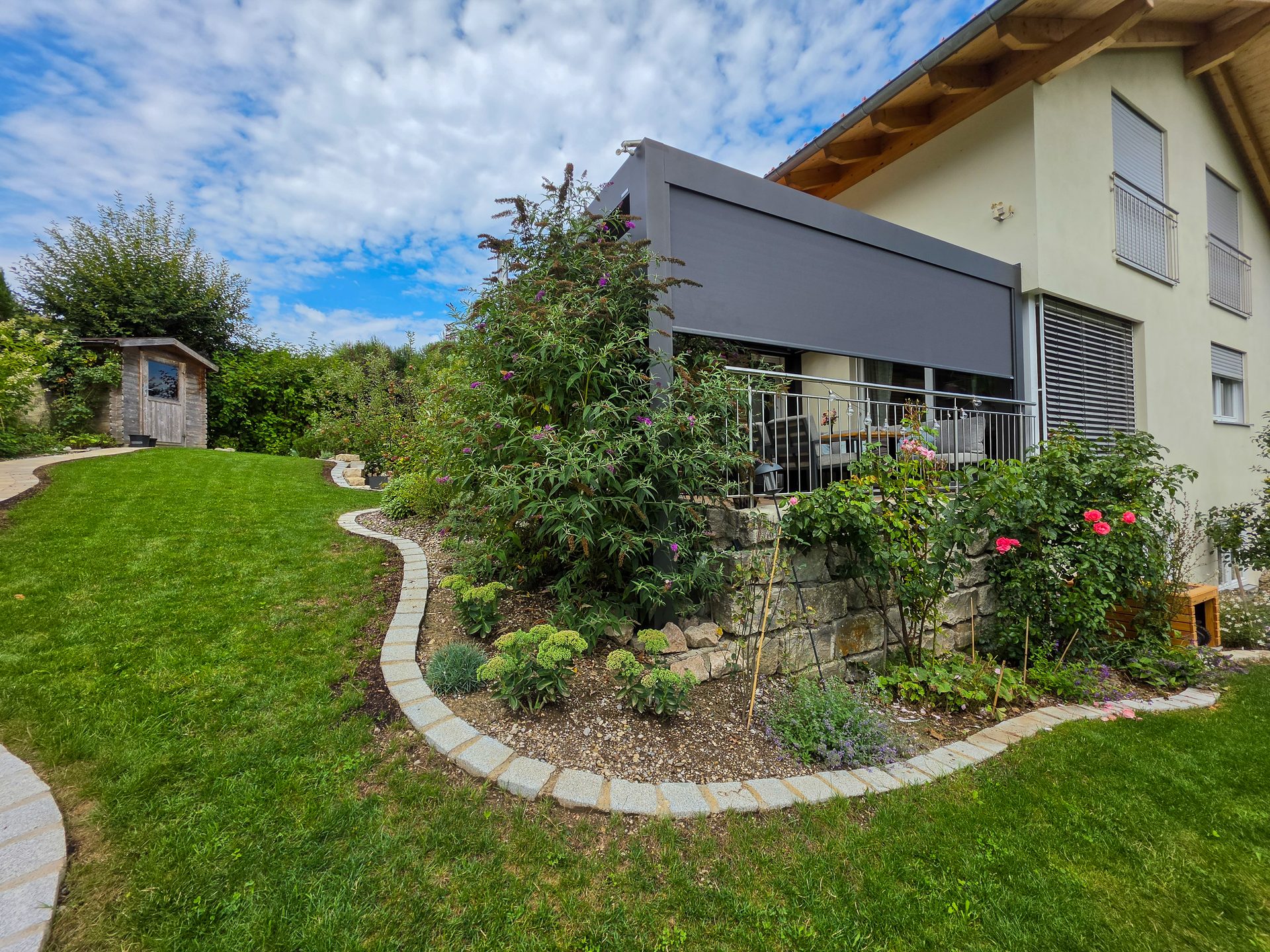 A modern house with a grey pergola over a balcony overlooking a lush garden with a shed and winding path.