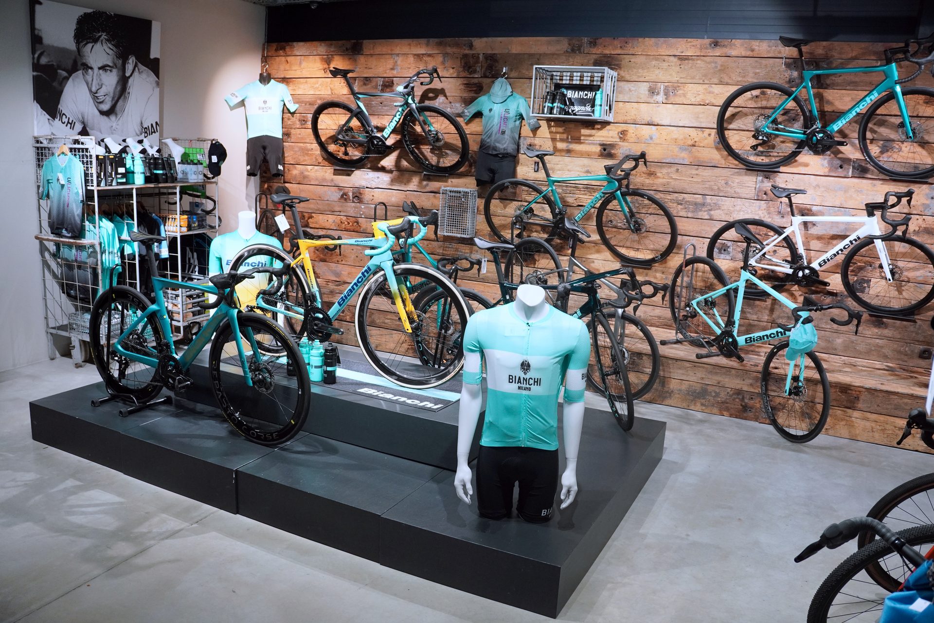 A bike shop interior displaying numerous Bianchi bicycles and cycling apparel on a wooden wall and black platforms.