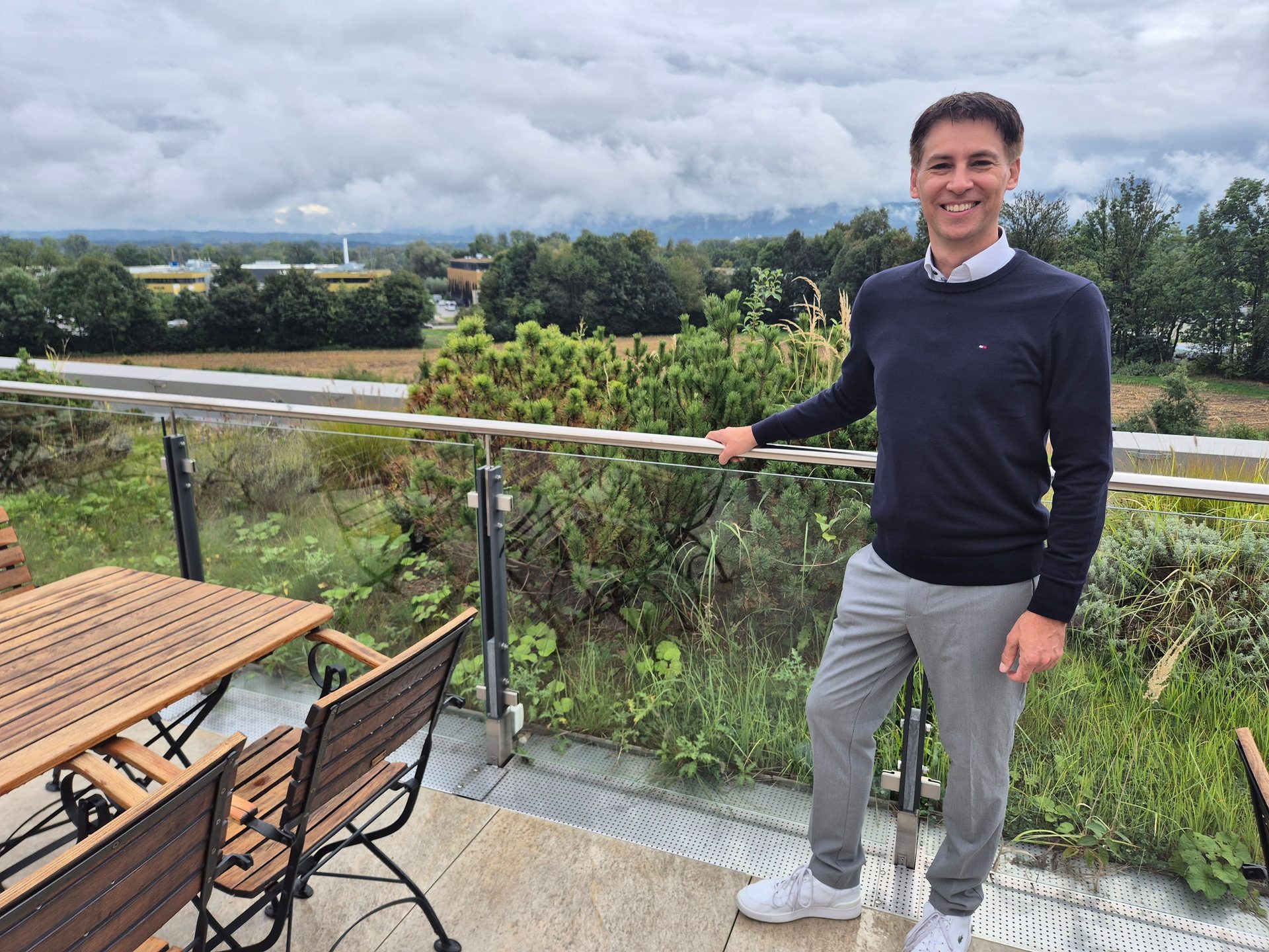 Smiling man on a balcony with a view of green trees and a cloudy sky.