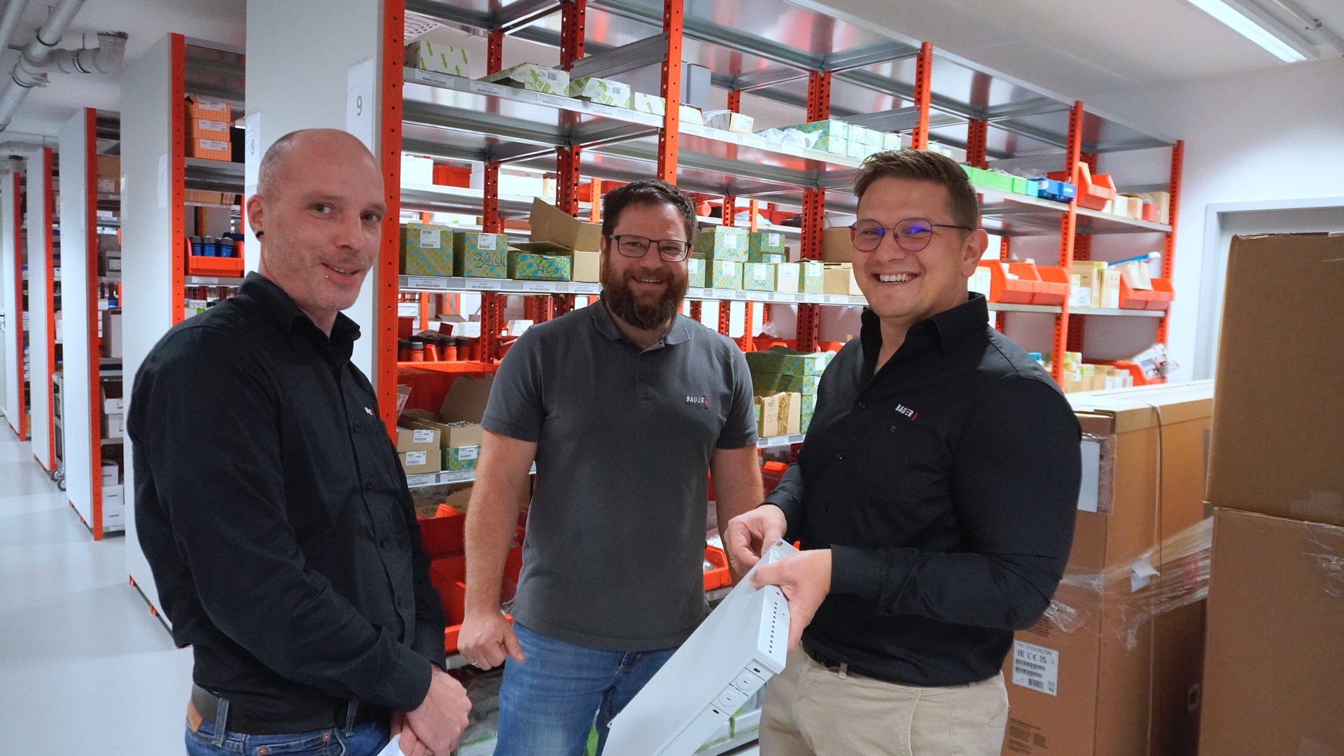 Three smiling men in a warehouse, one holding a white rectangular object.